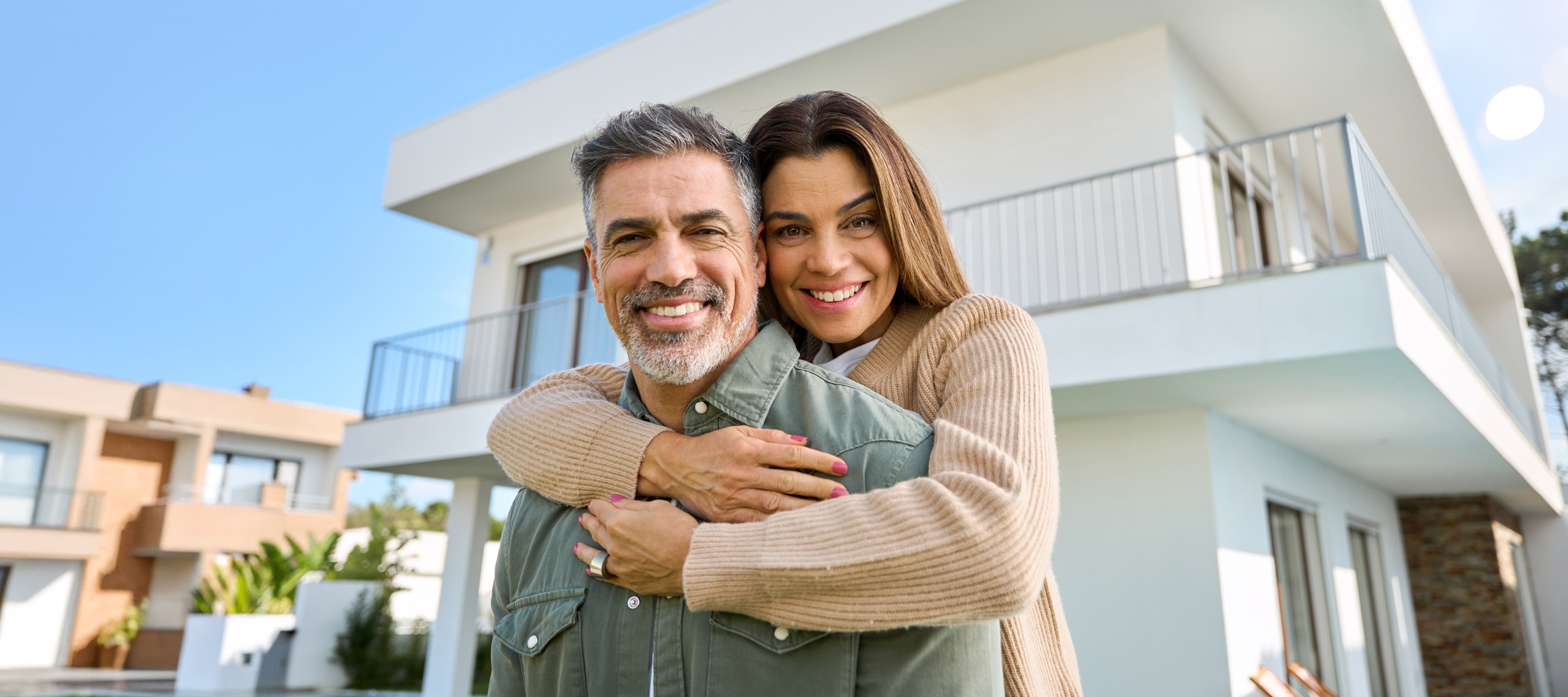 A man and woman embrace smiling at the camera in front of their newly purchased home