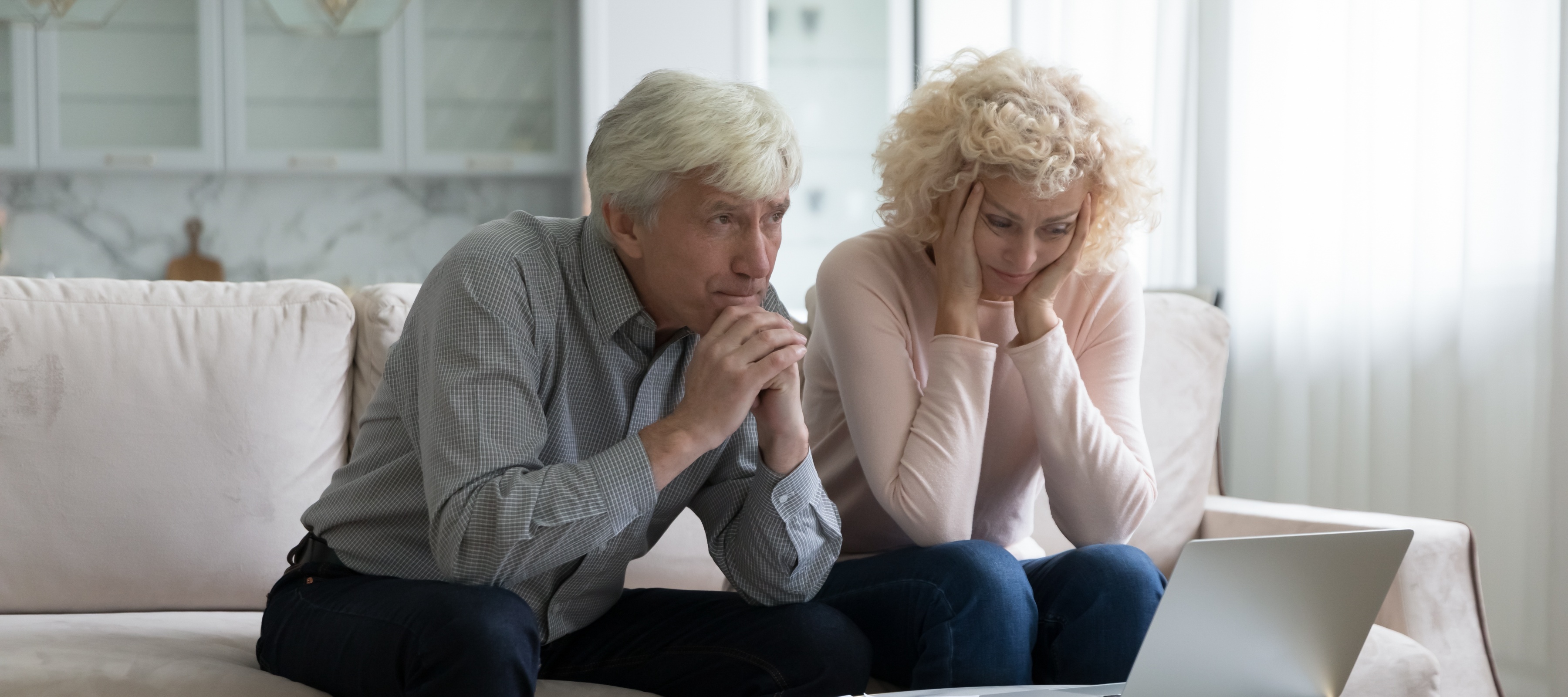 An older white man and woman sit next to each other on the couch looking stressed