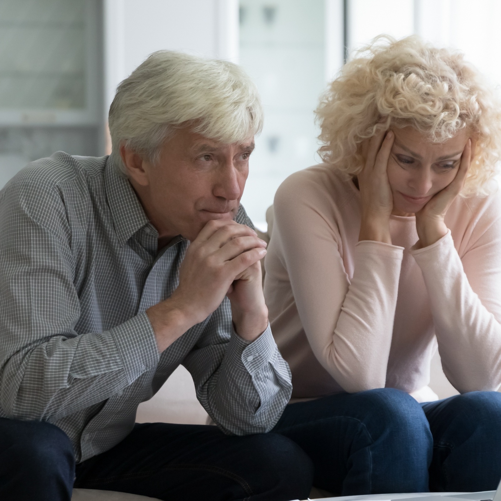 An older white man and woman sit next to each other on the couch looking stressed