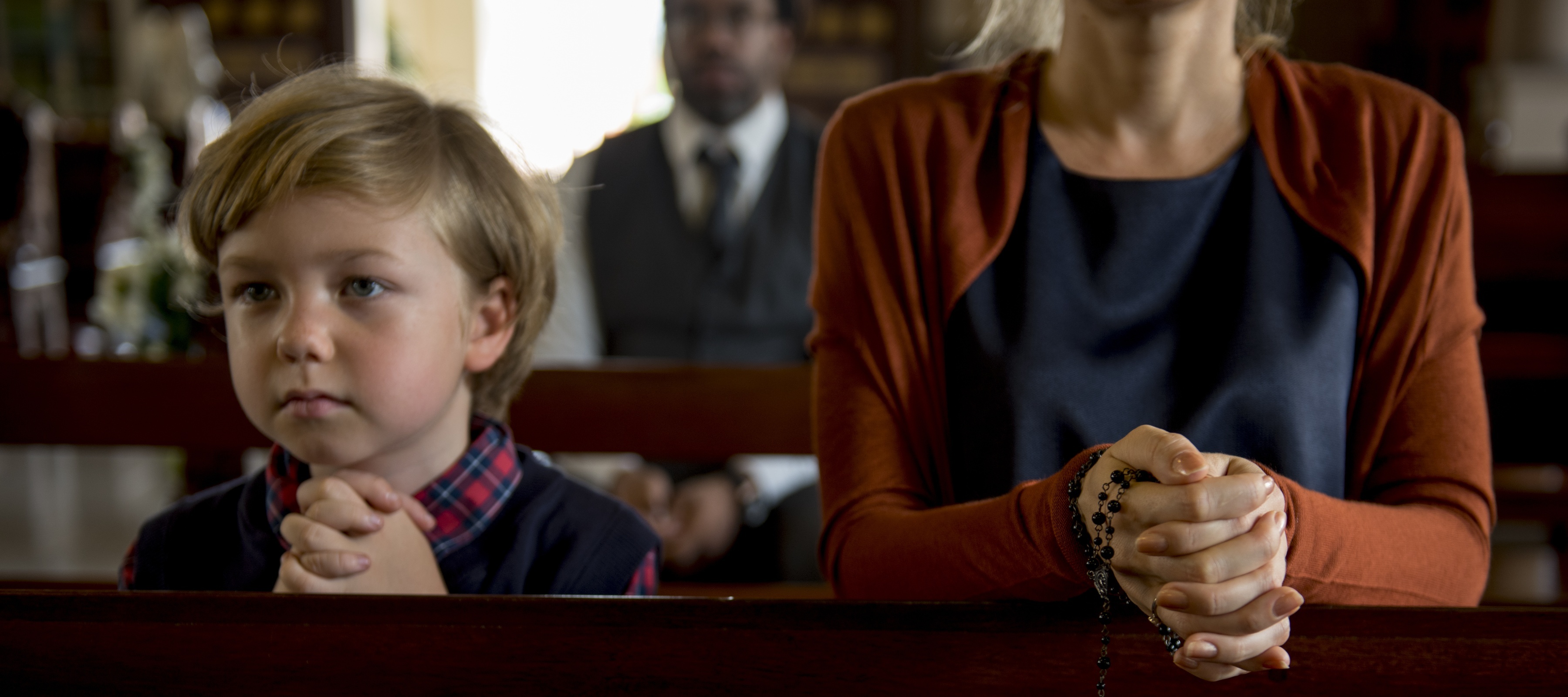 A sad-looking child and his mother clasp their hands at a pew in church