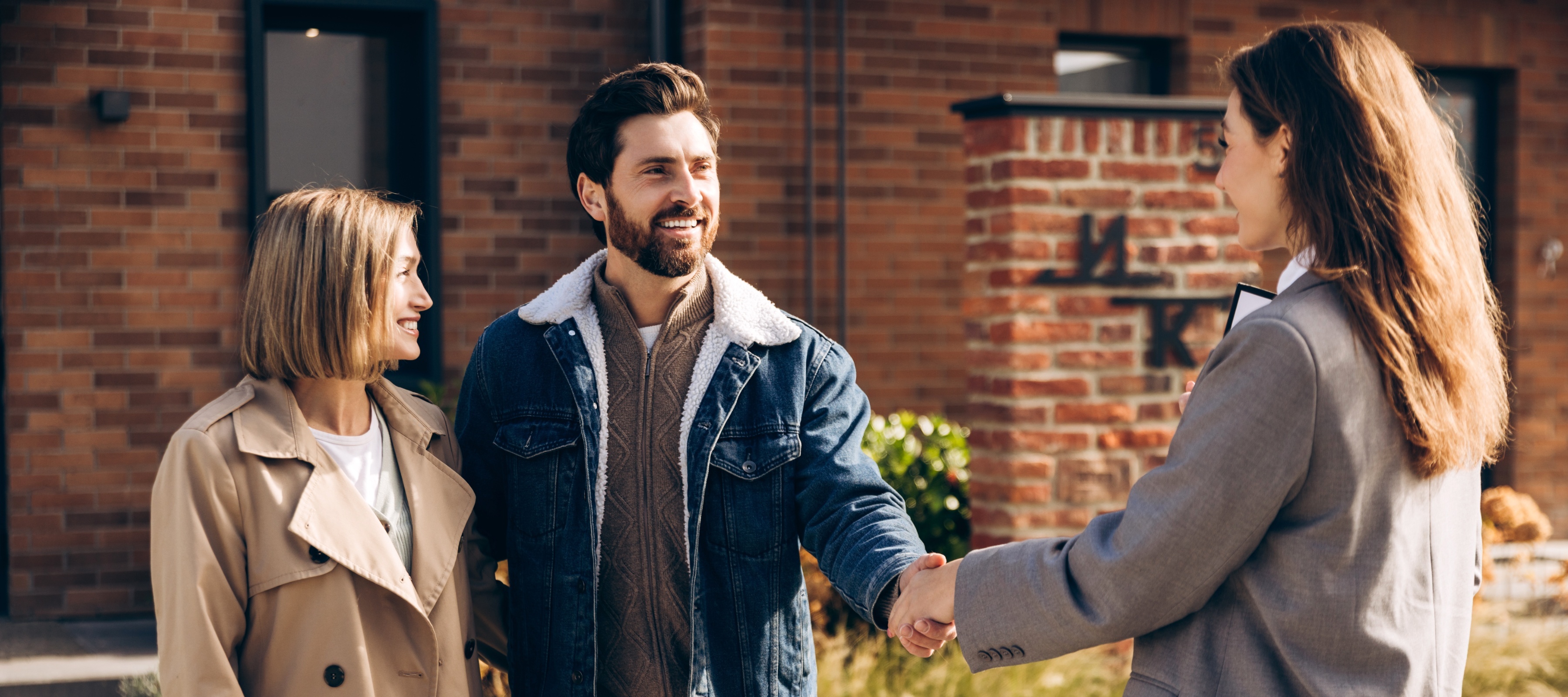 A man and woman accept keys from a real estate agent in front of a brick house