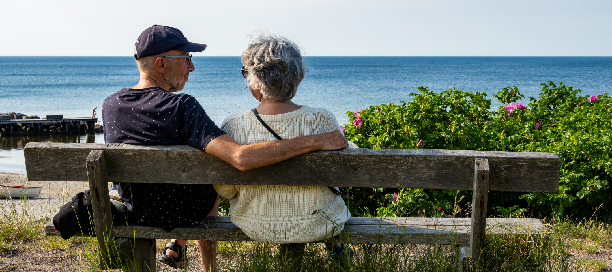 A senior couple look out to sea while sitting on a bench on the beach.