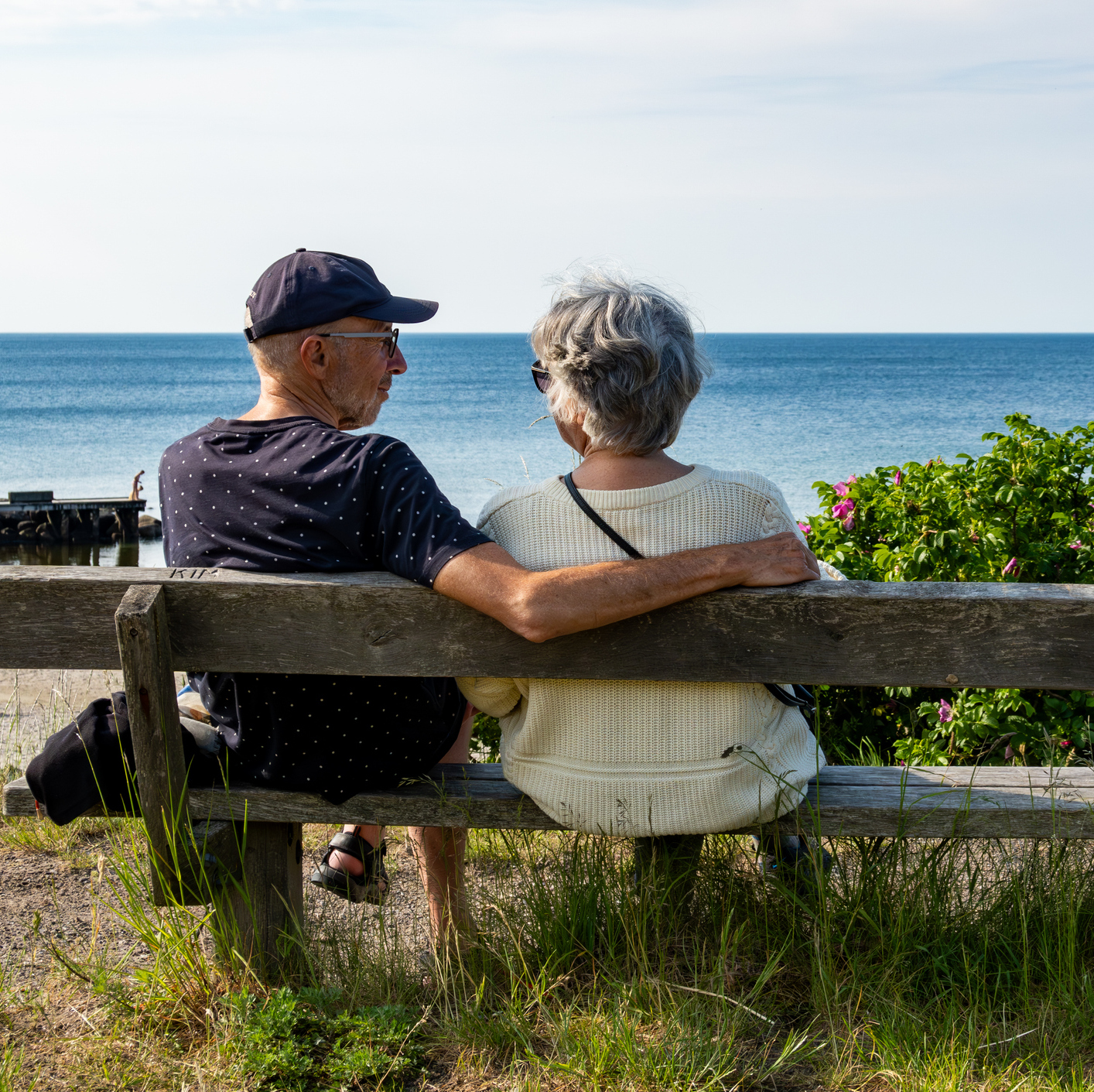 A senior couple look out to sea while sitting on a bench on the beach.