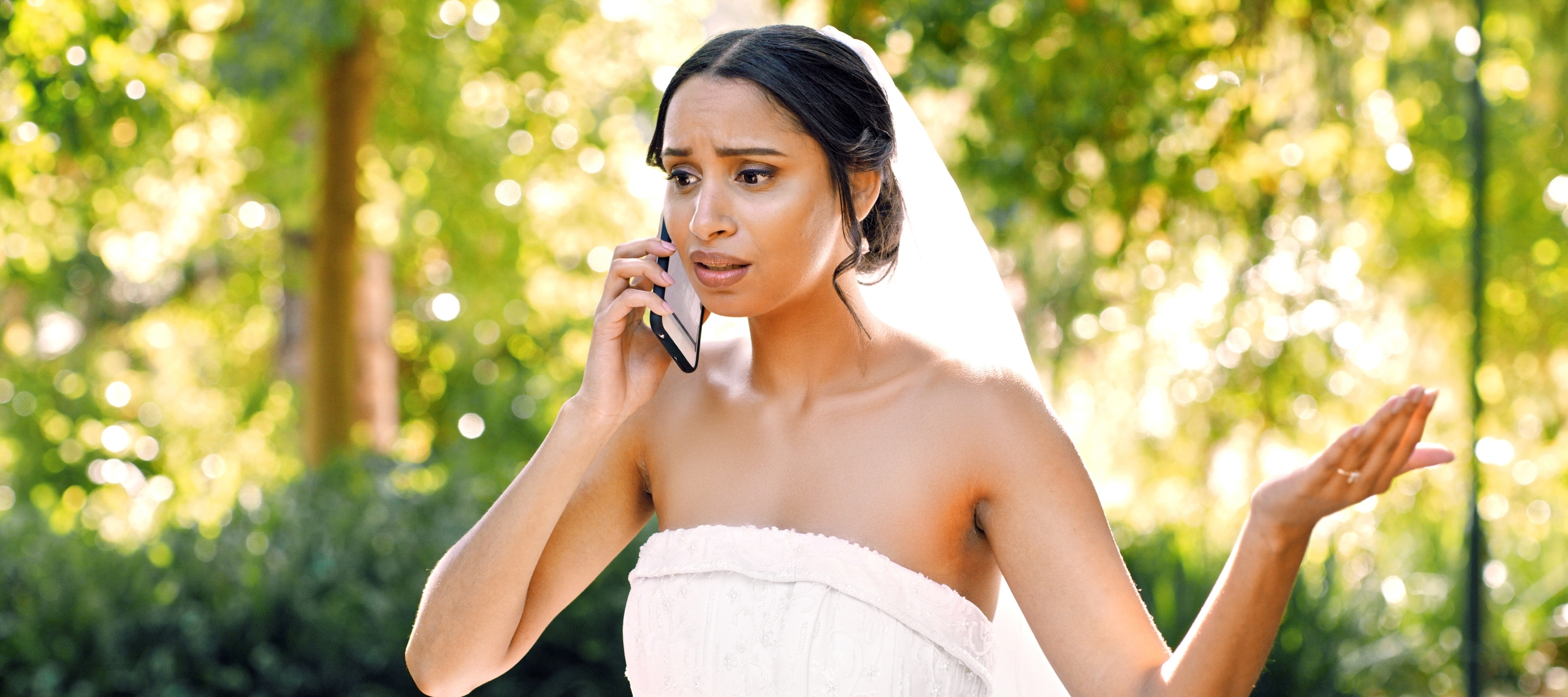 Woman in wedding gown stands outside in front of greenery while holding a phone to her ear, looking worried and upset.
