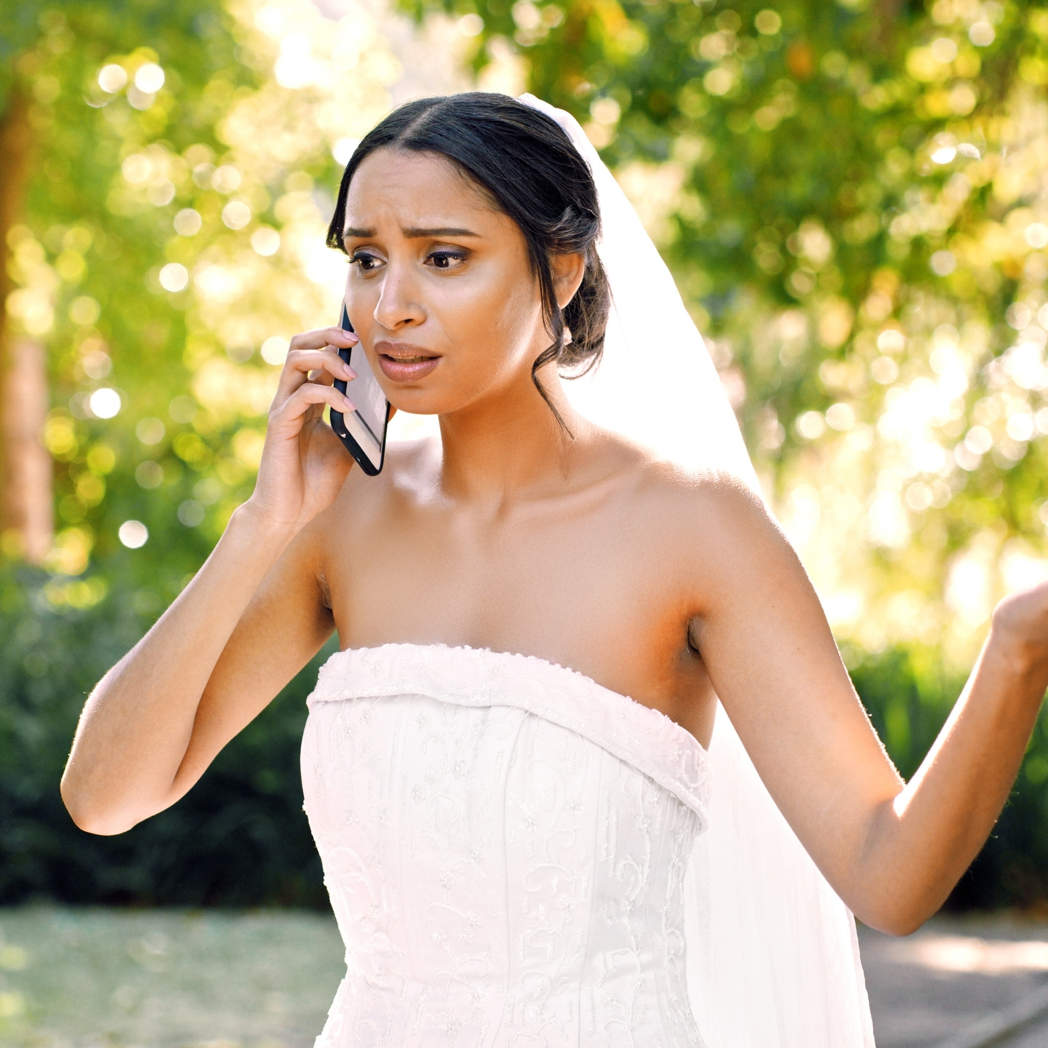 Woman in wedding gown stands outside in front of greenery while holding a phone to her ear, looking worried and upset.
