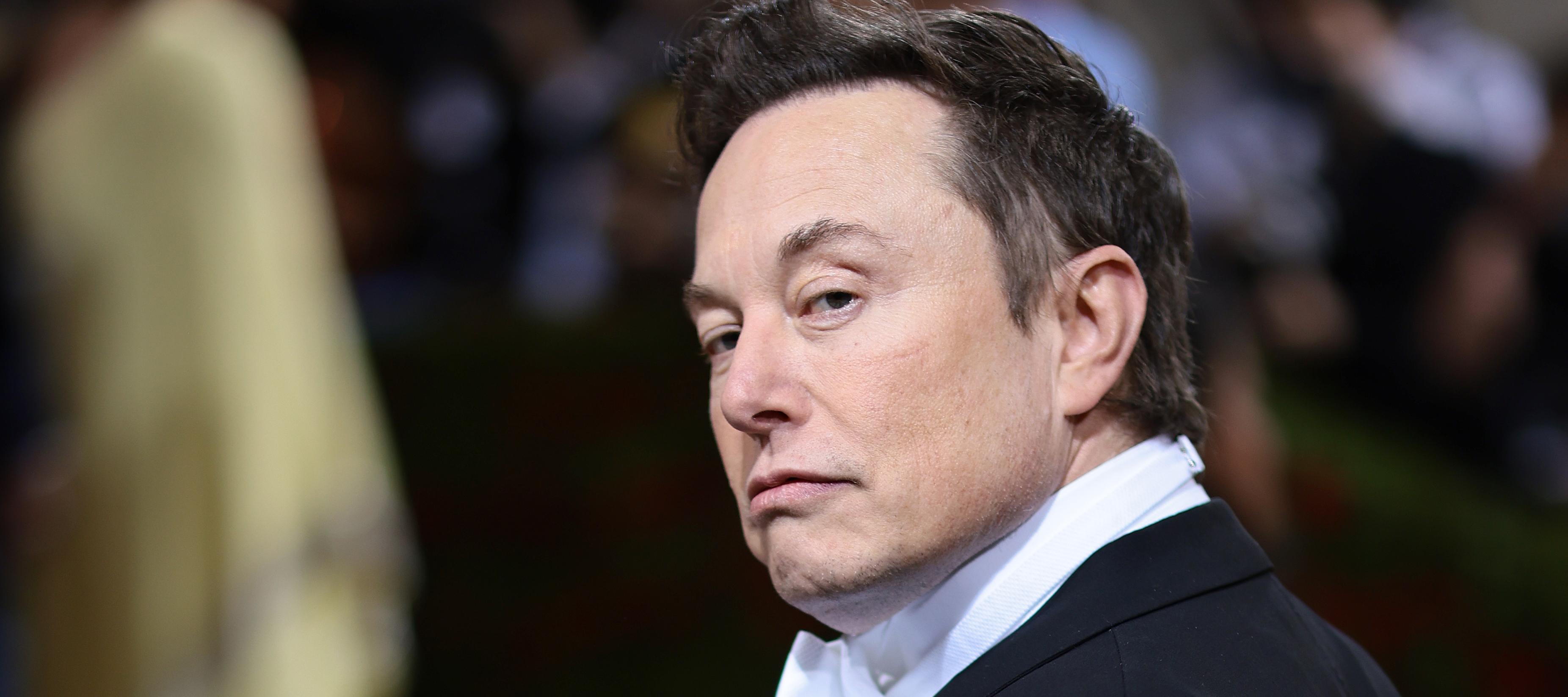 Elon Musk glances over one shoulder, dressed in a black suit and clean white shirt.