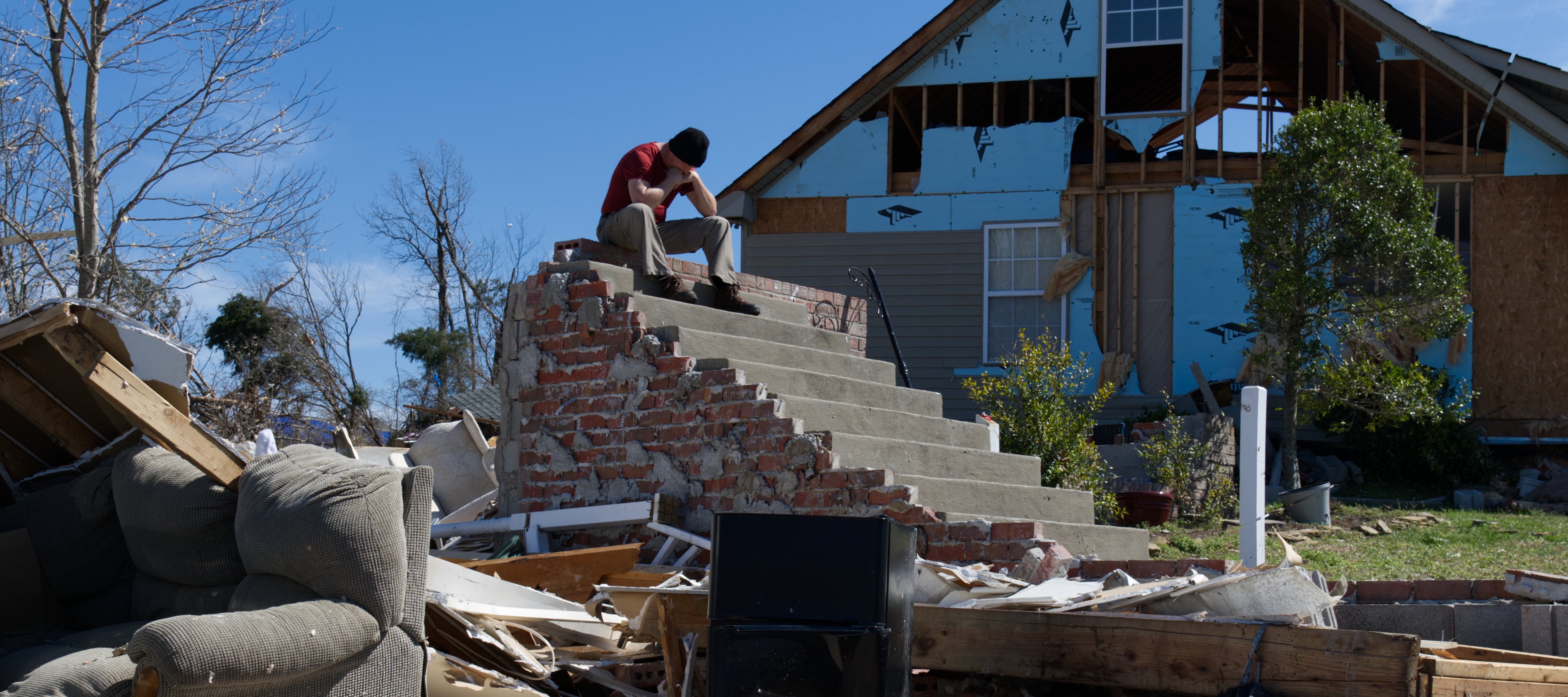 A man sits atop steps that remain from his home that was destroyed by a tornado. He holds his head in his hands.