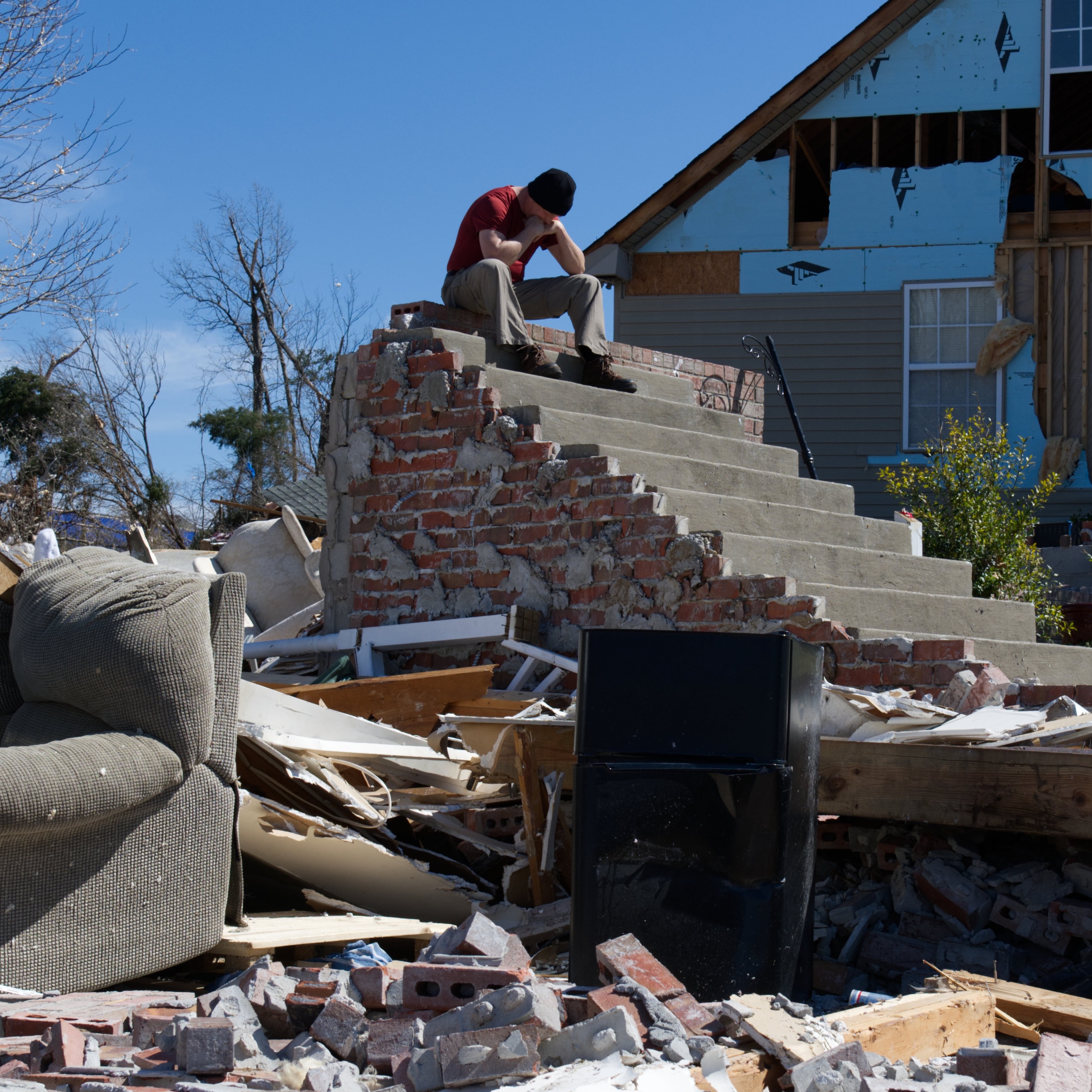A man sits atop steps that remain from his home that was destroyed by a tornado. He holds his head in his hands.