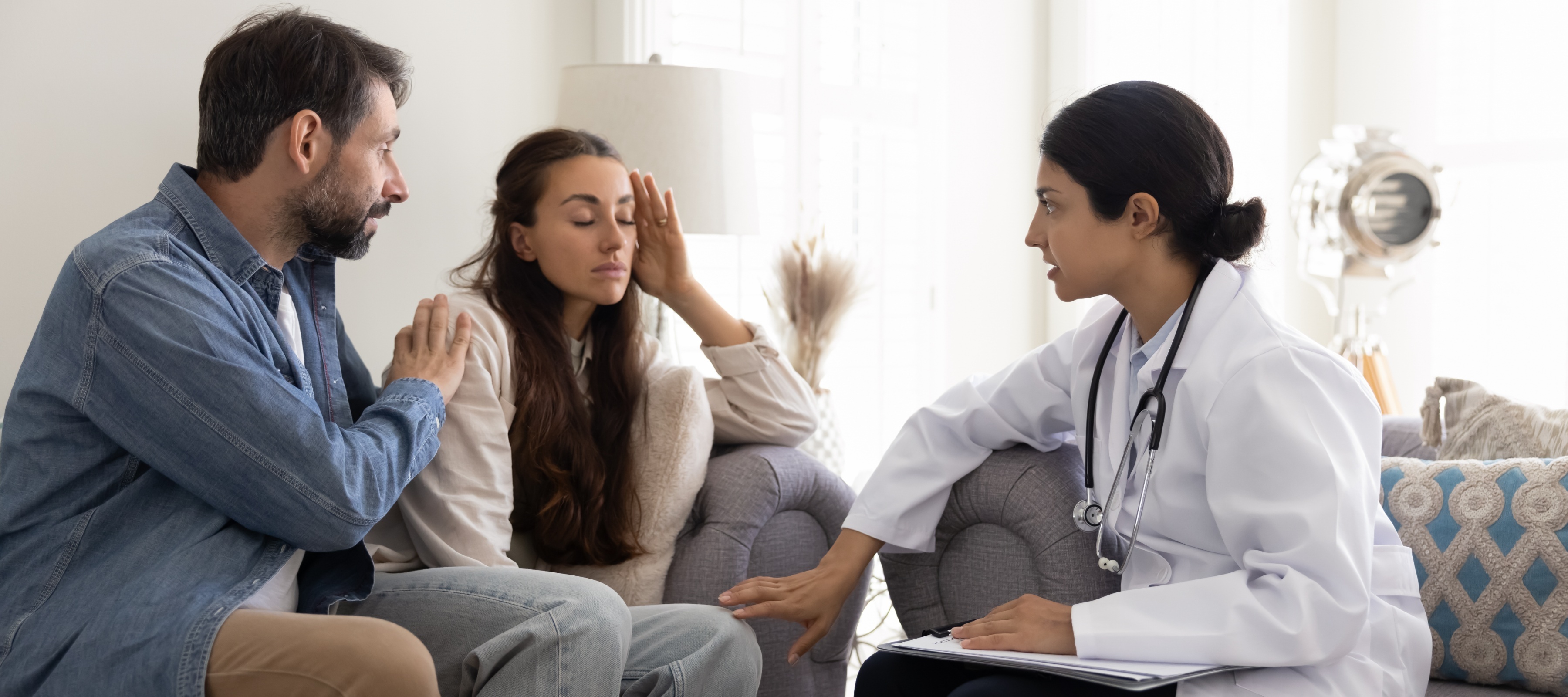 A man and a doctor console a worried-looking woman inside a doctor's office