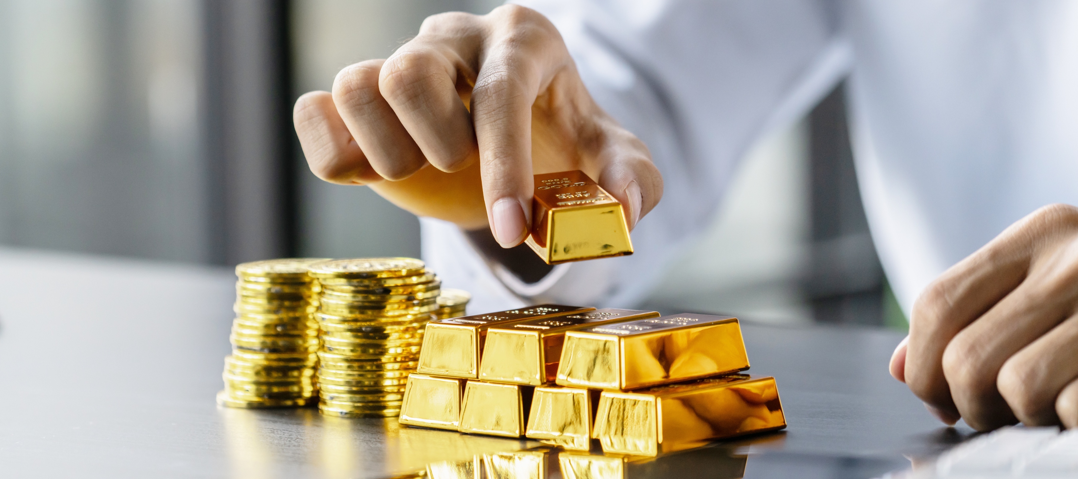A hand stacking gold bullion and gold coins on a table