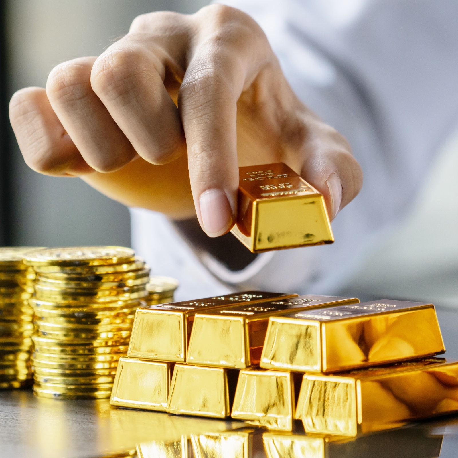 A hand stacking gold bullion and gold coins on a table