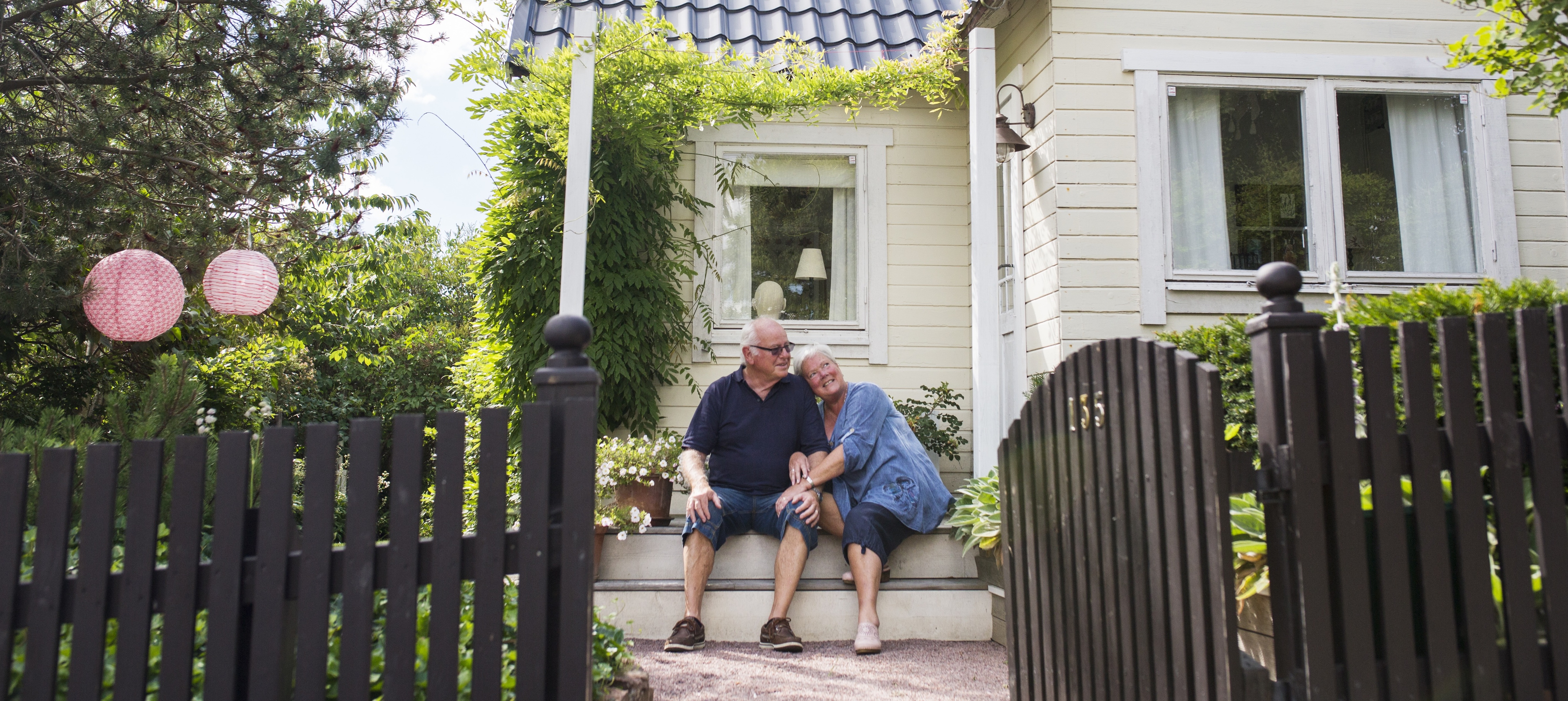 An older man and woman sit together on the porch of their house in a suburban environment.