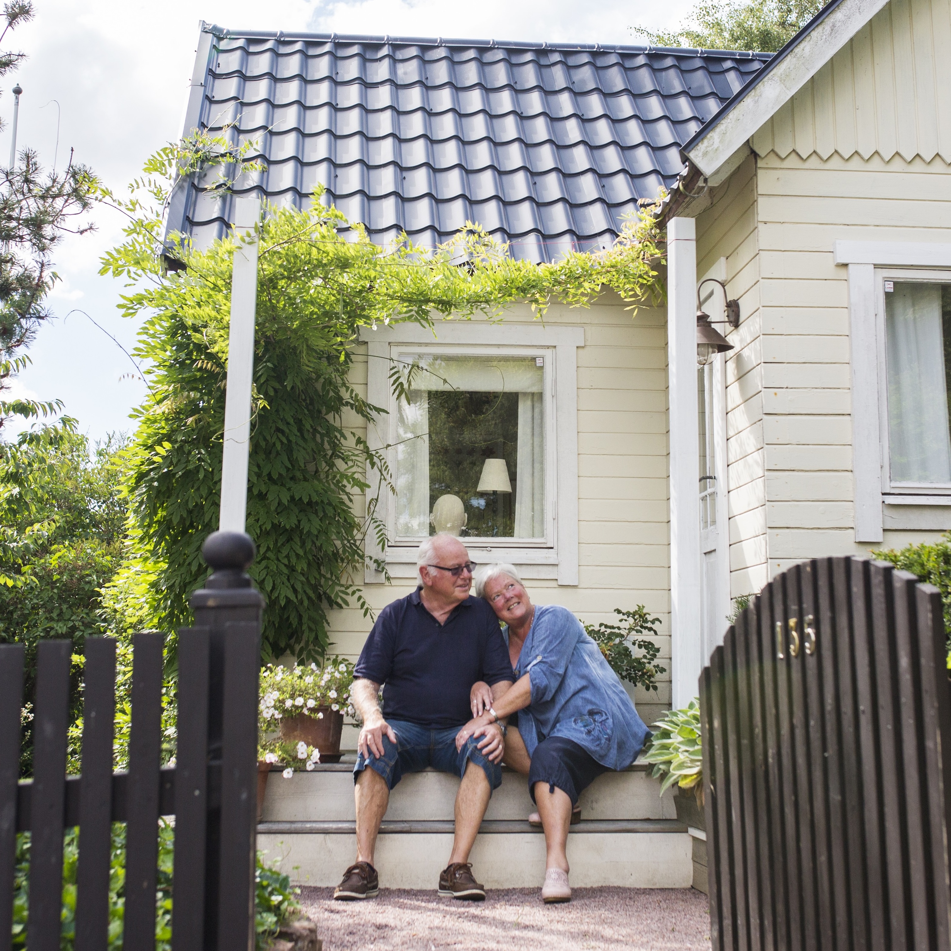 An older man and woman sit together on the porch of their house in a suburban environment.