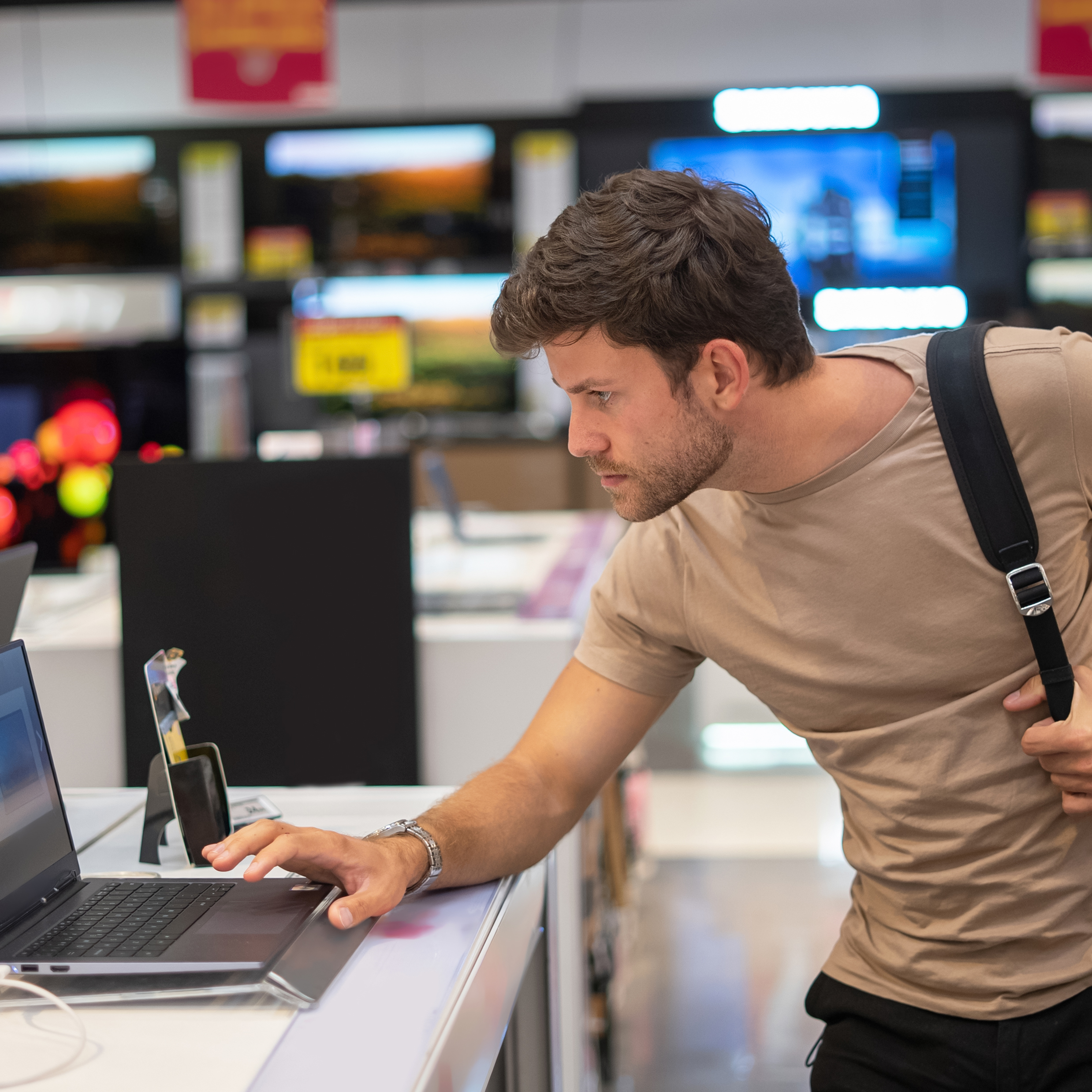 A man stands in a brightly lit electronics store, leaning slightly to look at a laptop computer on display.