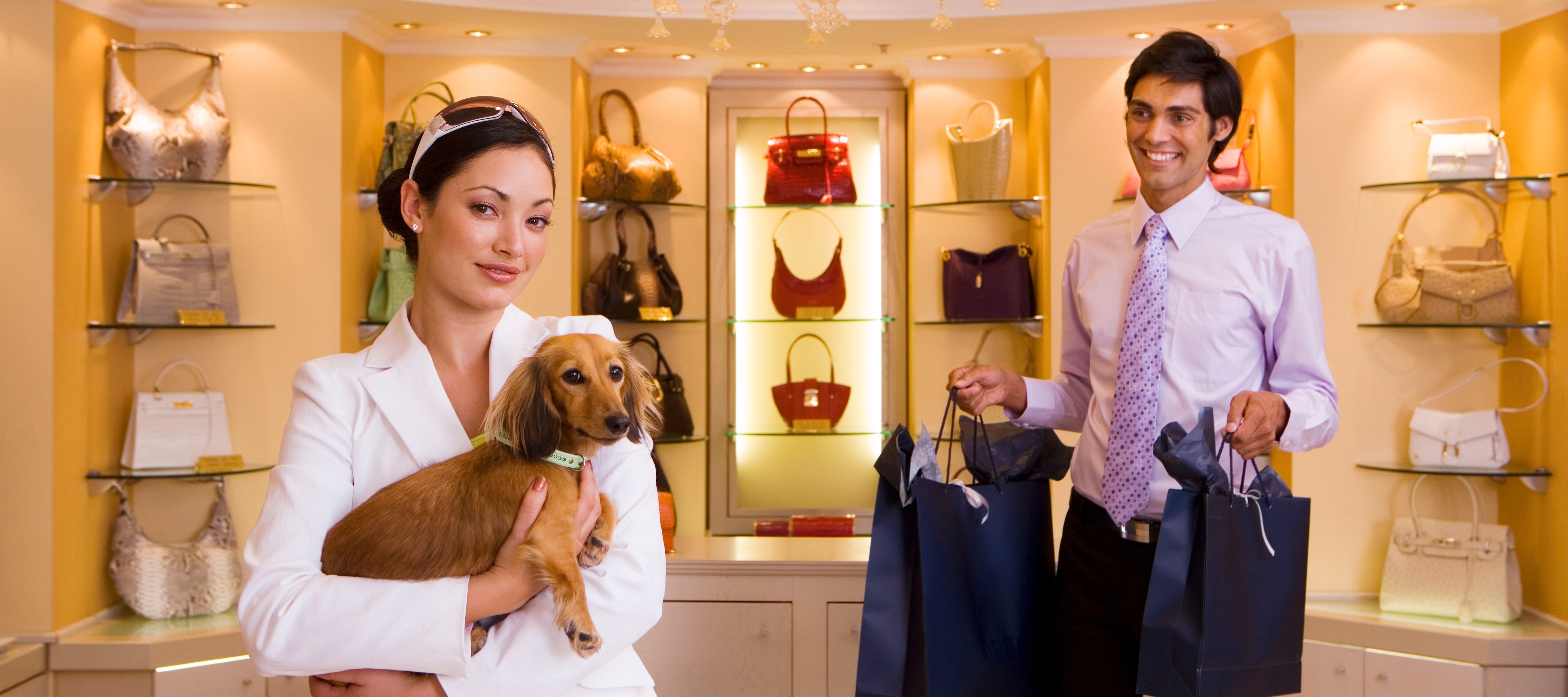 A smartly dressed woman holds a brown dachshund while standing in an elegant handbag store next to a man carrying shopping bags.