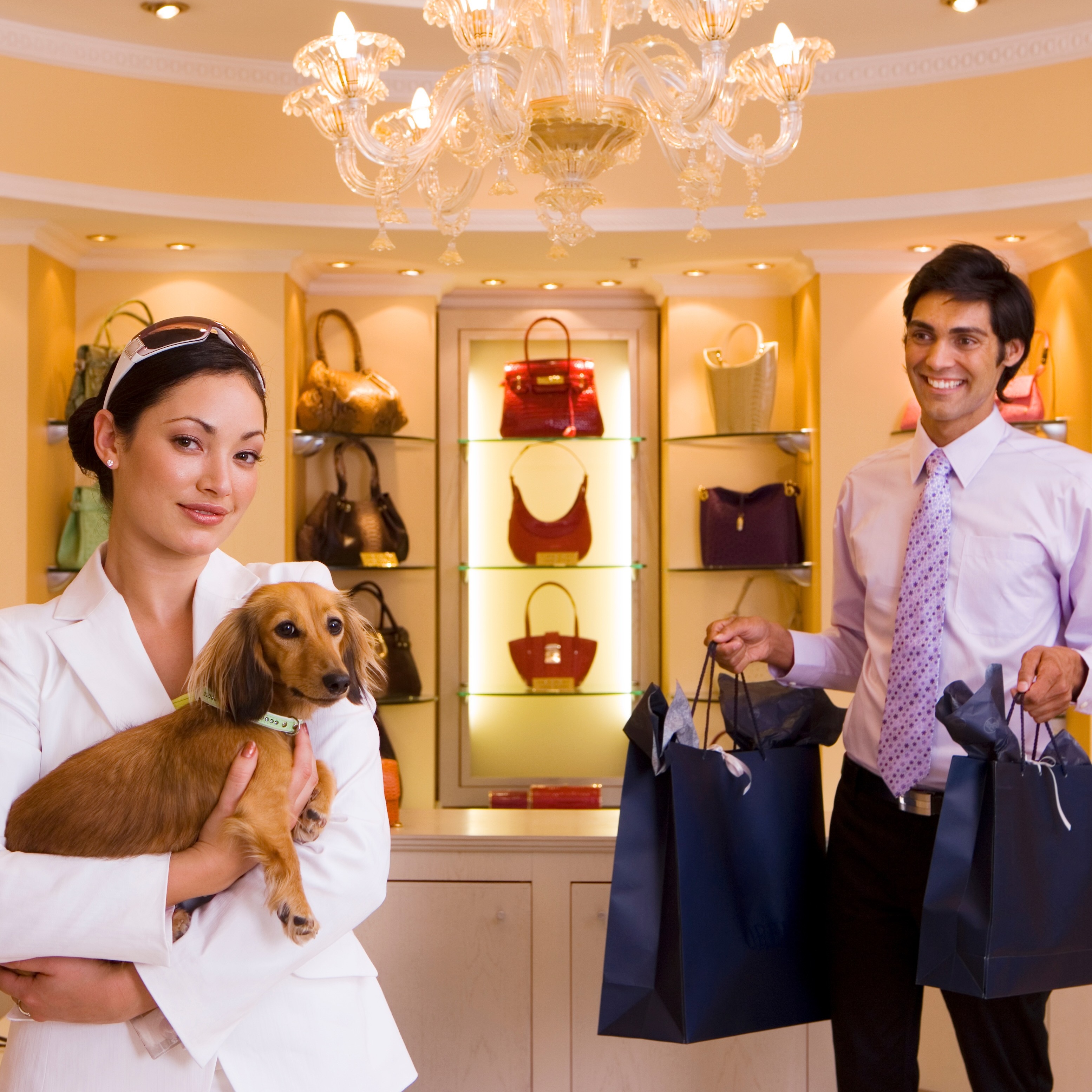 A smartly dressed woman holds a brown dachshund while standing in an elegant handbag store next to a man carrying shopping bags.