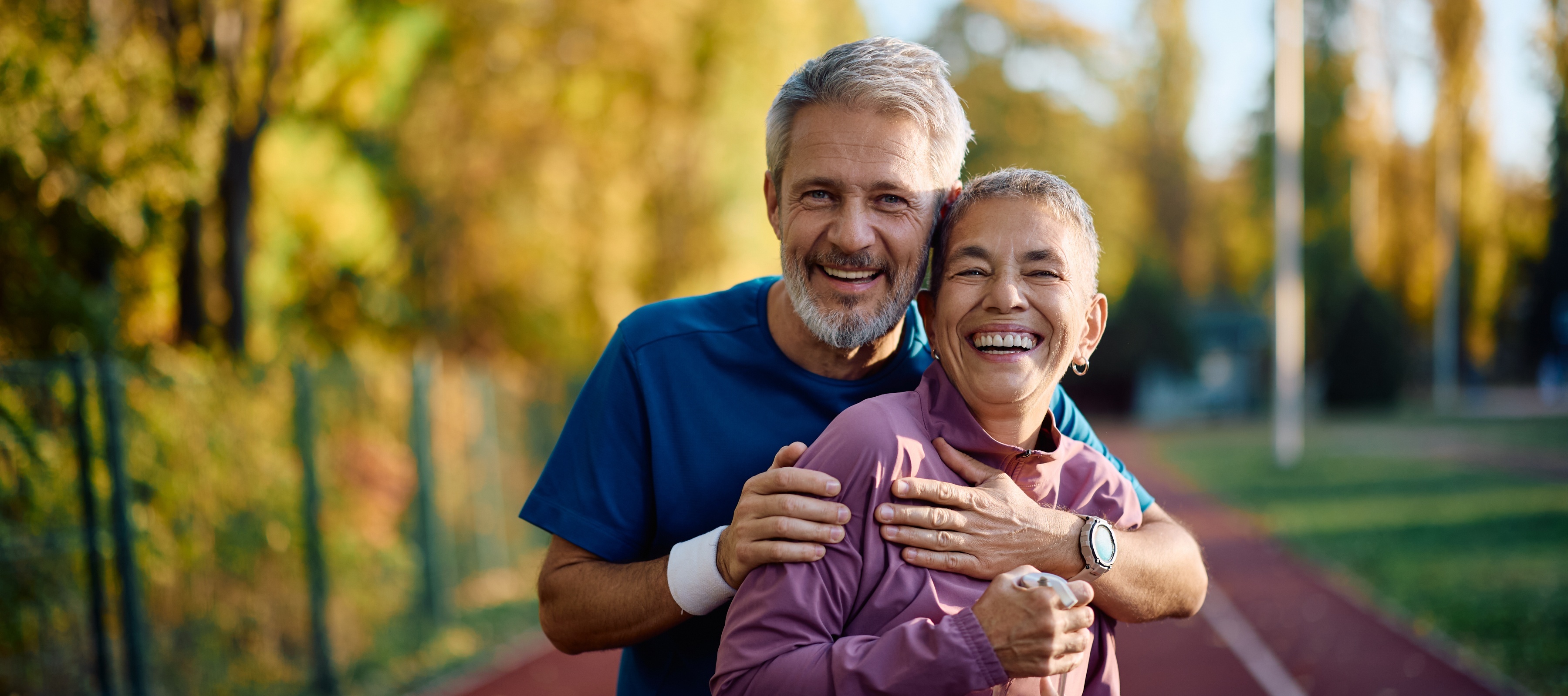 Older couple smile for the camera, seen posing together in workout clothes at an outdoor track.