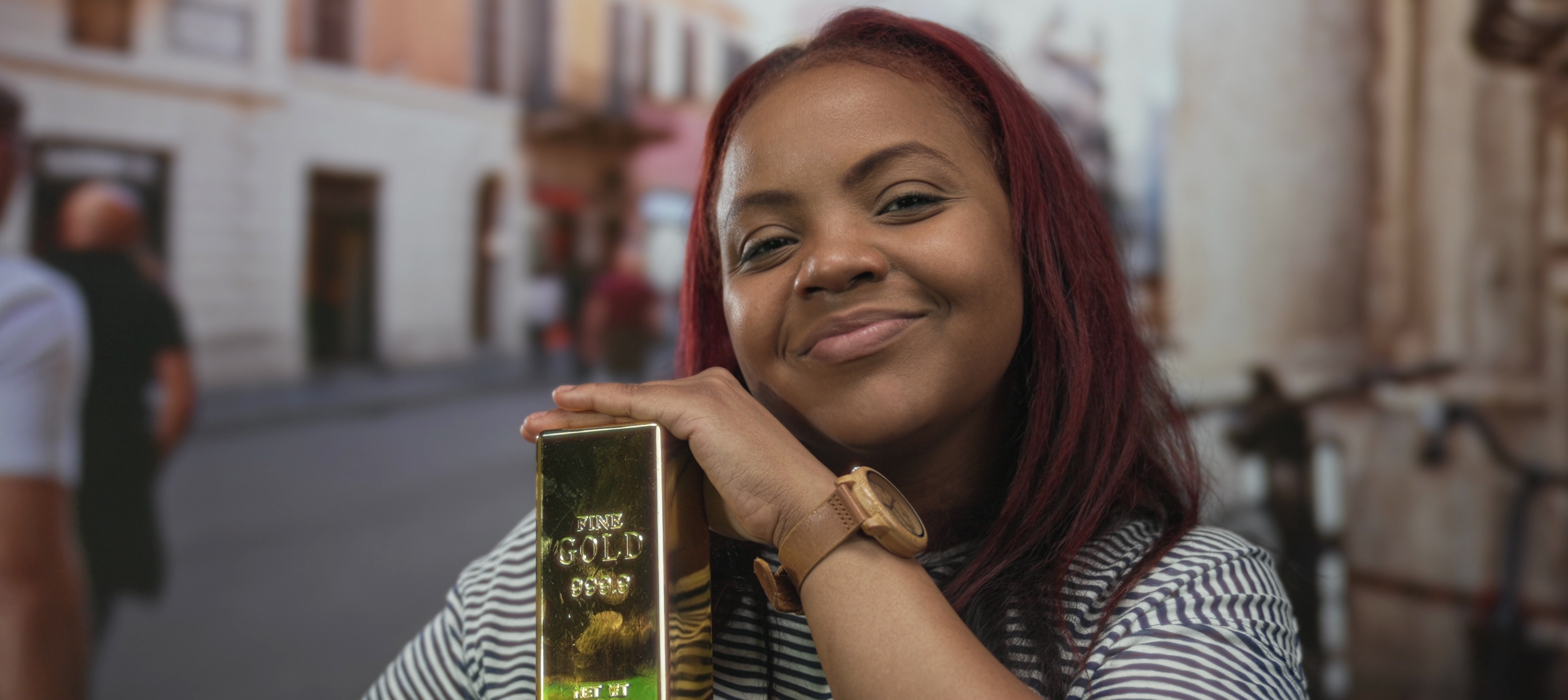 A woman holds a gold bar with both hands on busy crowded city street; she is happy and smiling broadly.