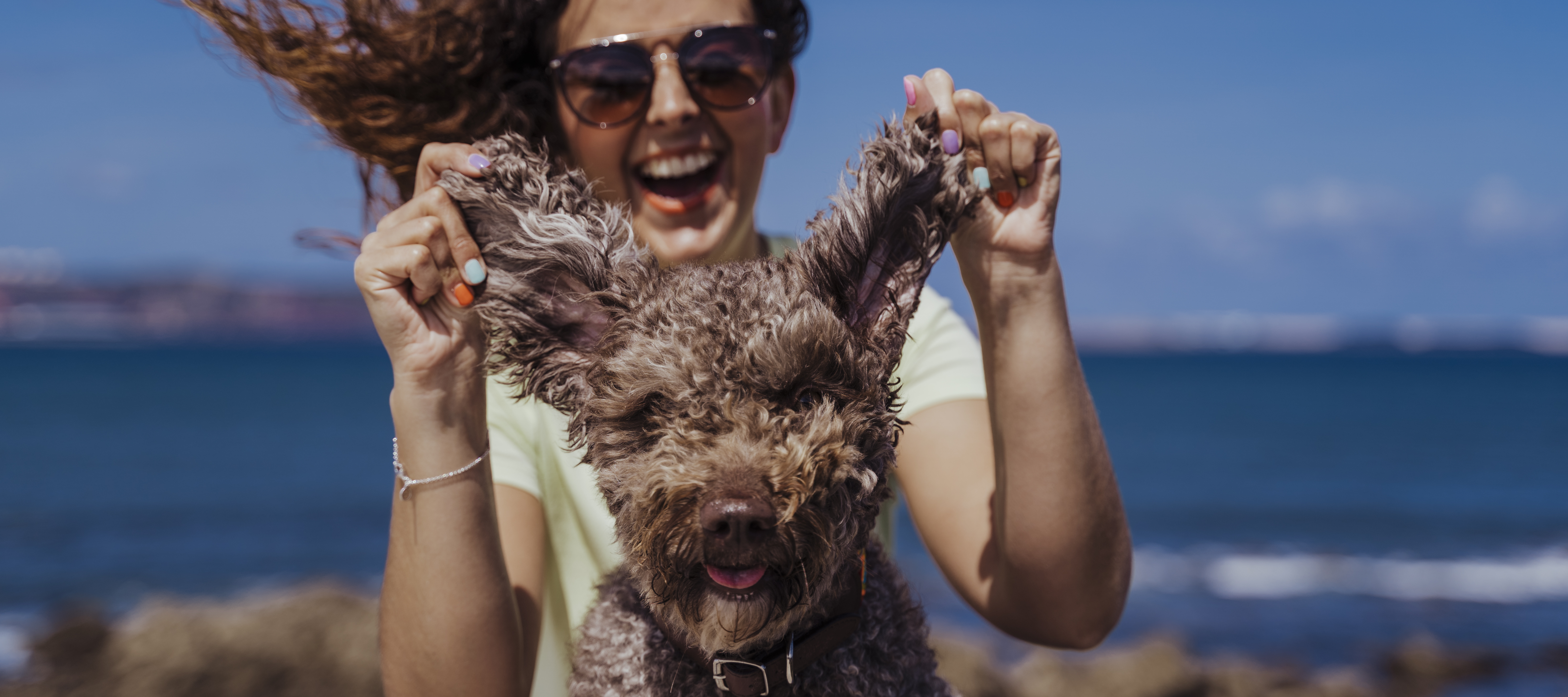 A smiling woman with curly hair and sunglasses playfully holds up the ears of her fluffy brown dog.