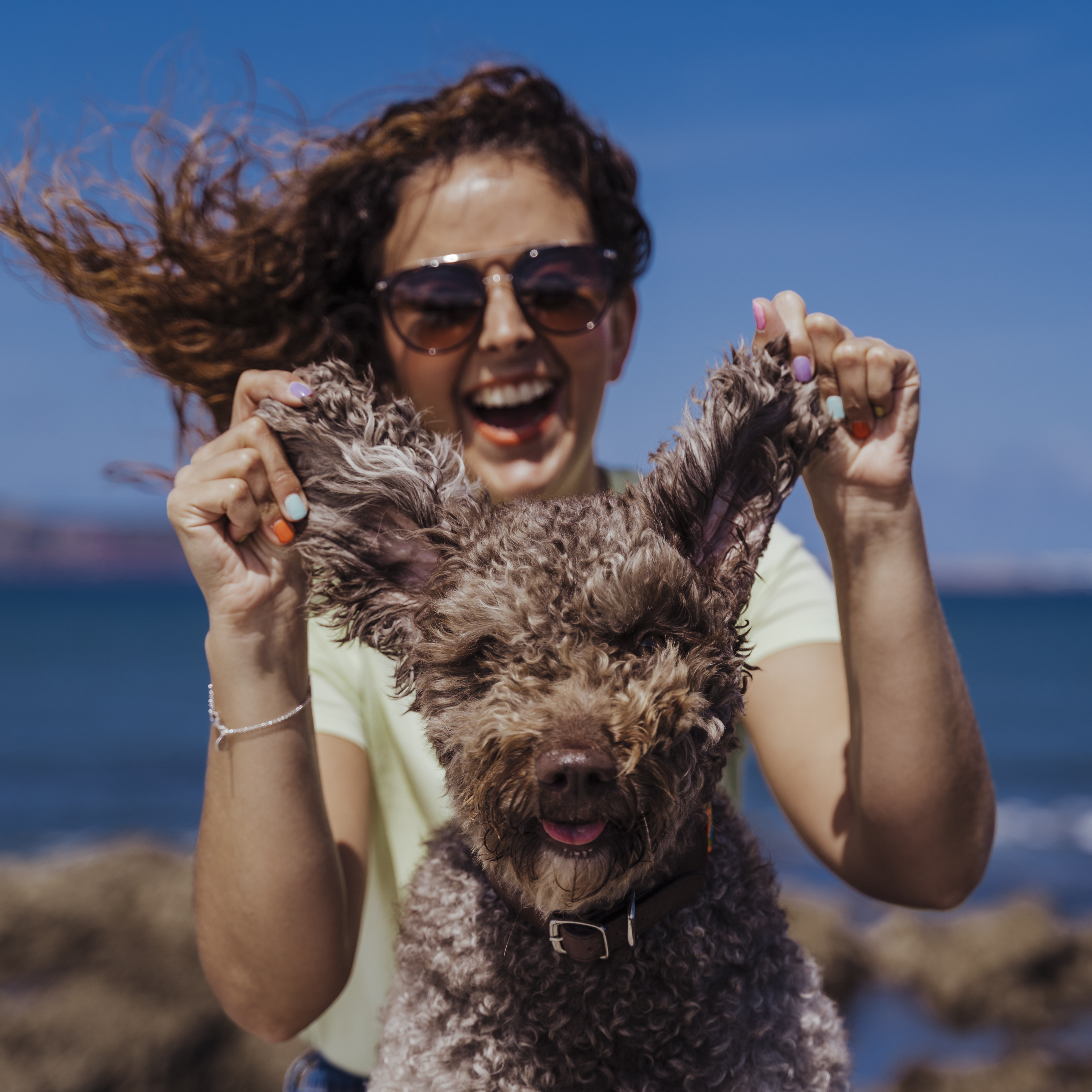A smiling woman with curly hair and sunglasses playfully holds up the ears of her fluffy brown dog.