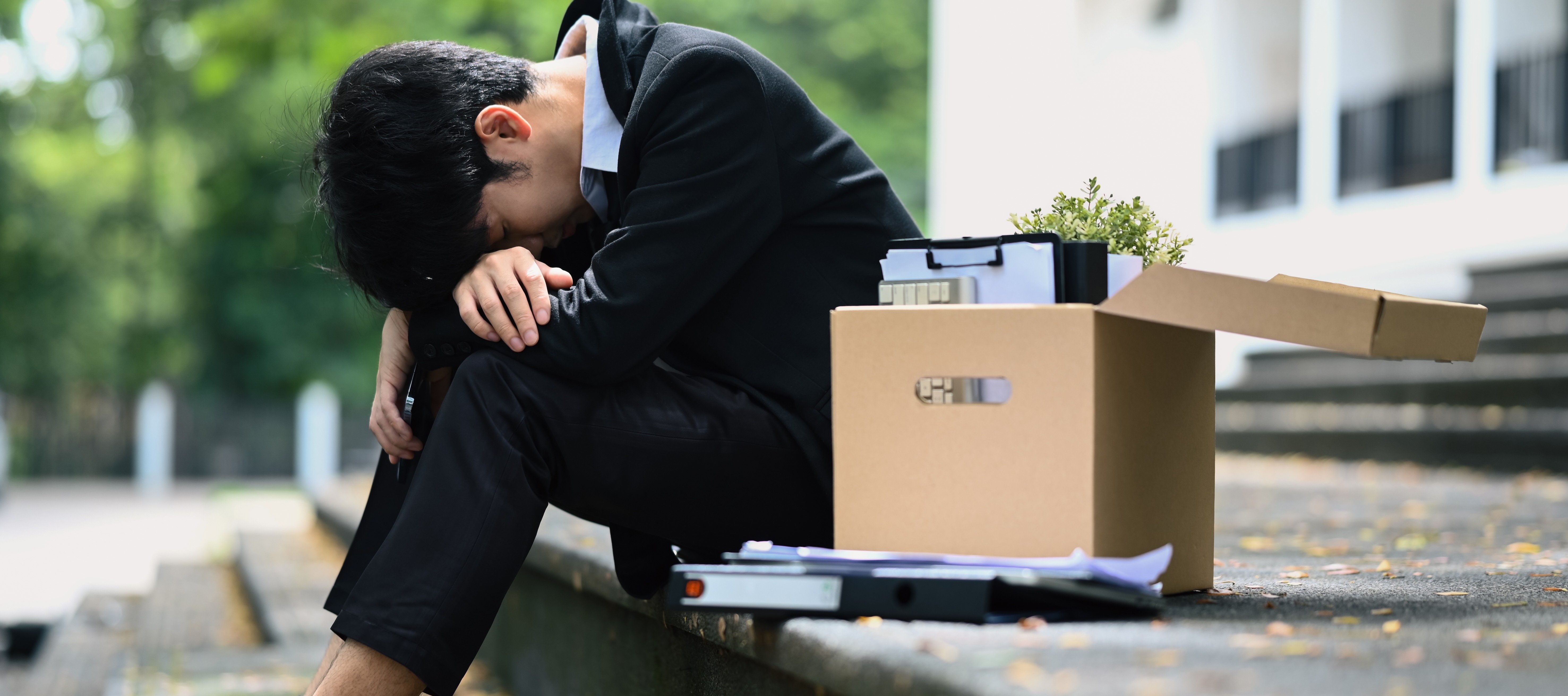 A young man in a suit sits on a concrete staircase outside with his head in his hands.