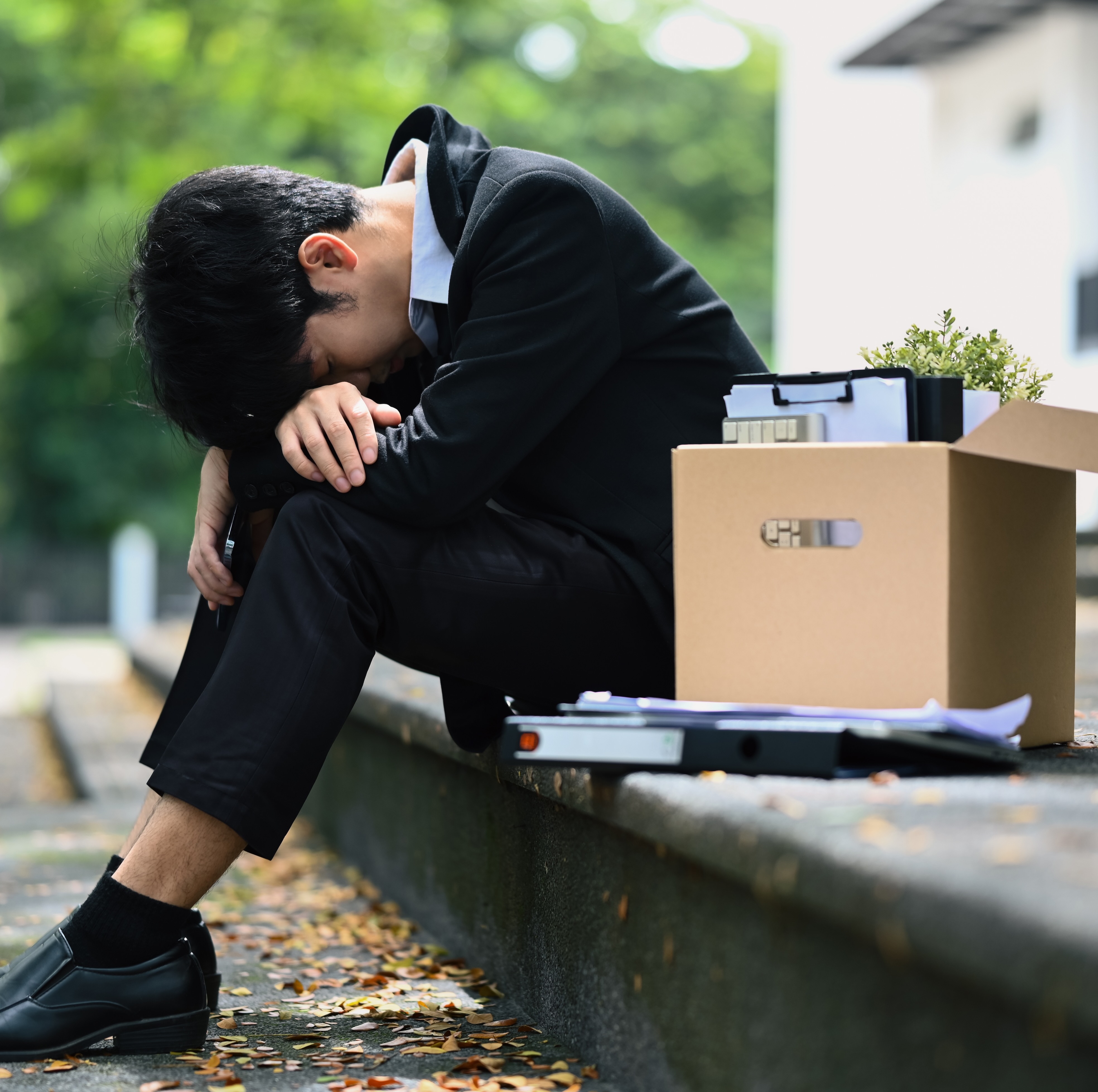 A young man in a suit sits on a concrete staircase outside with his head in his hands.