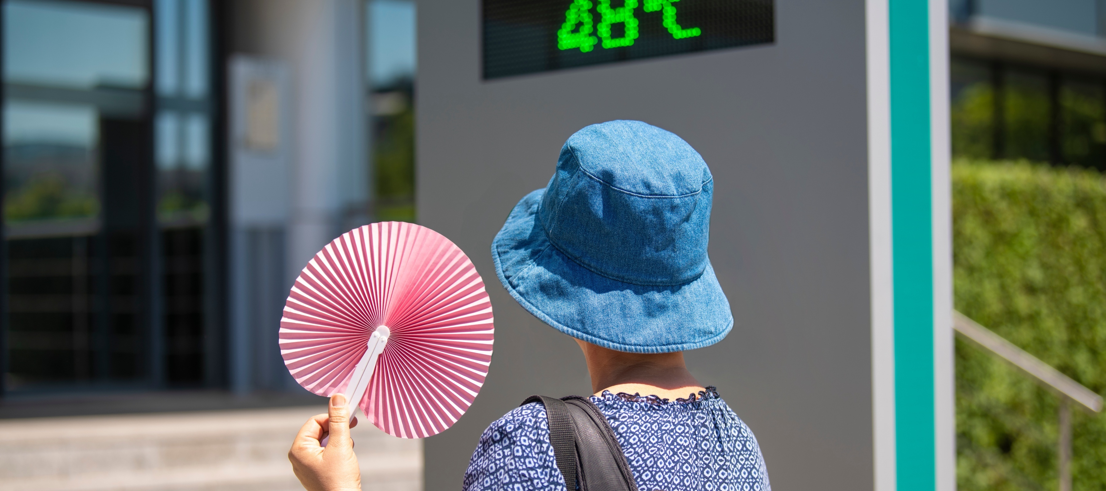 An old woman in a bucket hat holds a fan in front of a temperature reading during a heatwave