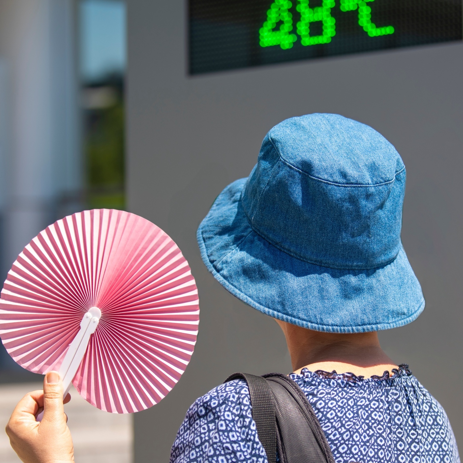 An old woman in a bucket hat holds a fan in front of a temperature reading during a heatwave