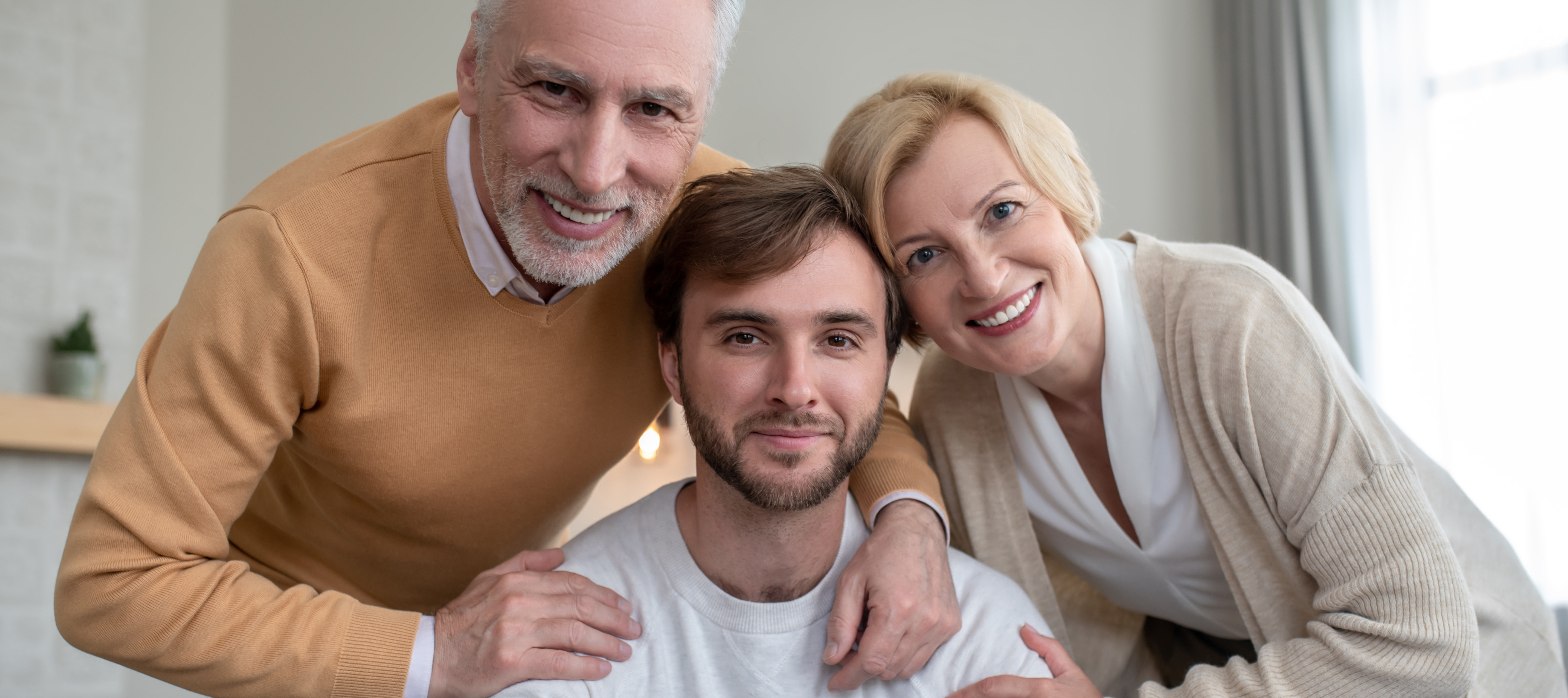 A younger adult with brown hair sits at a table while his parents place their hands on his shoulders, embracing him.