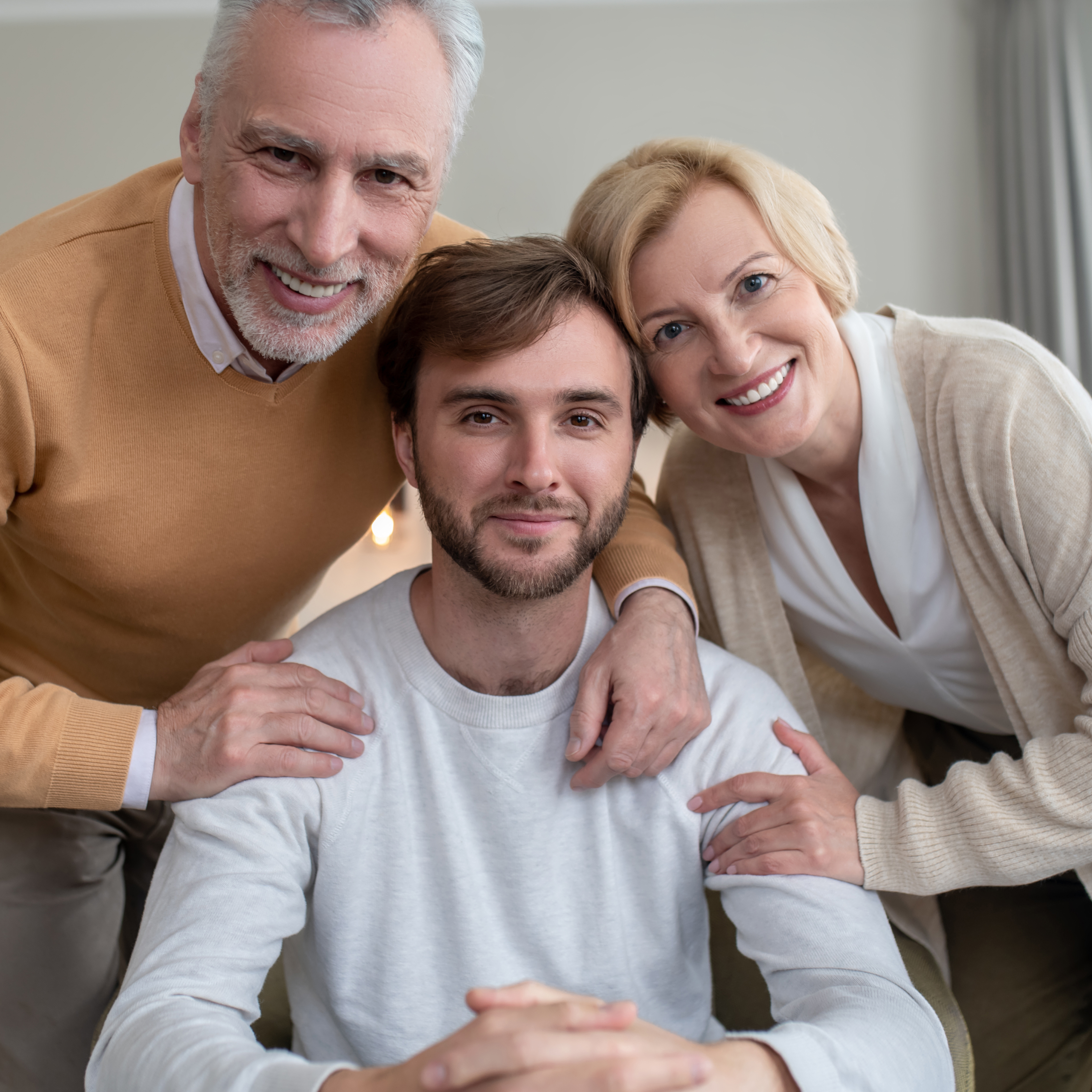 A younger adult with brown hair sits at a table while his parents place their hands on his shoulders, embracing him.