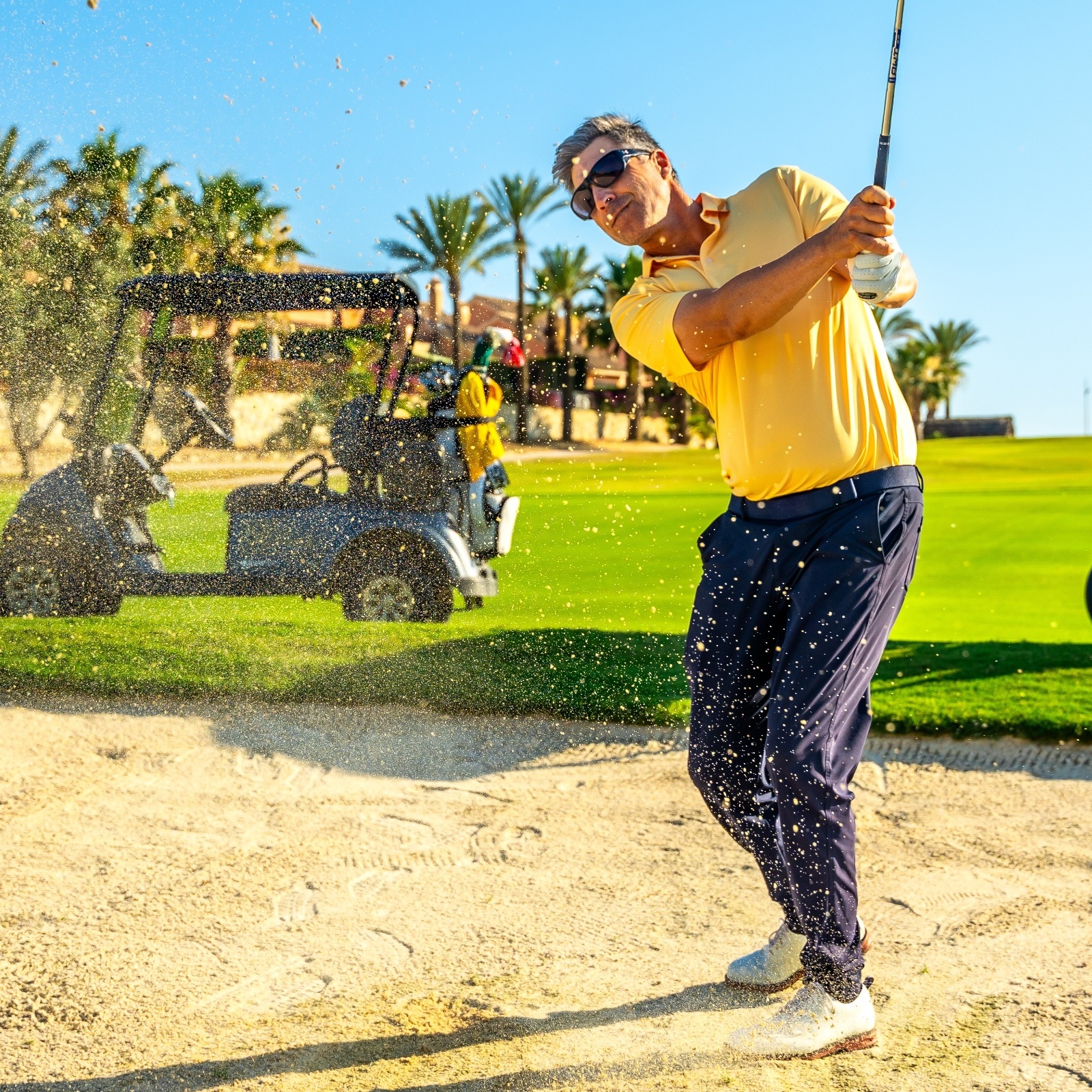 Male golfer hitting ball out of sand trap with golf cart and other players in background on sunny day.