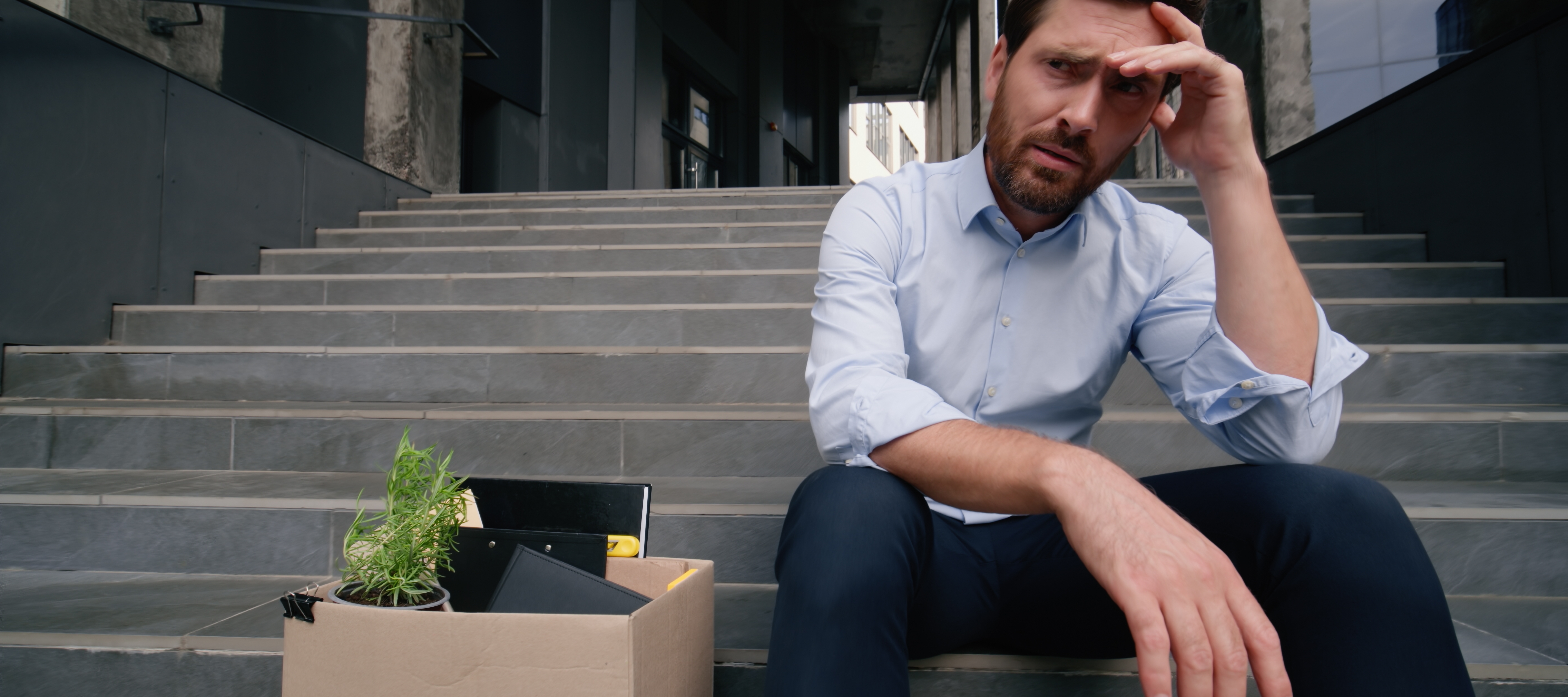 A man sits on the steps outside of his office building with a pensive look and a box of his belongings beside him.