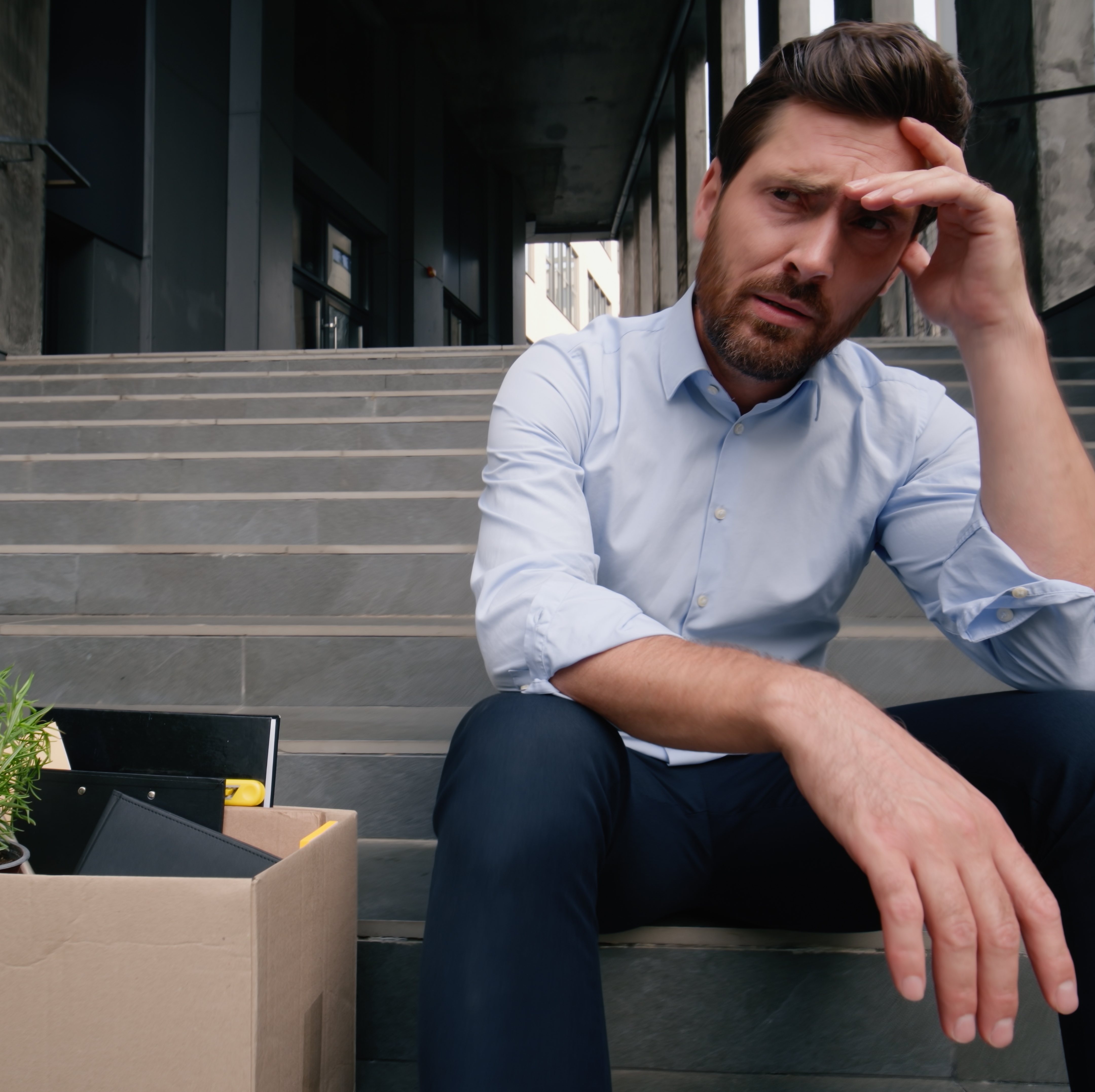 A man sits on the steps outside of his office building with a pensive look and a box of his belongings beside him.