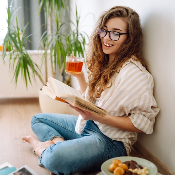 Young woman sitting on floor, drinking tea and reading book.