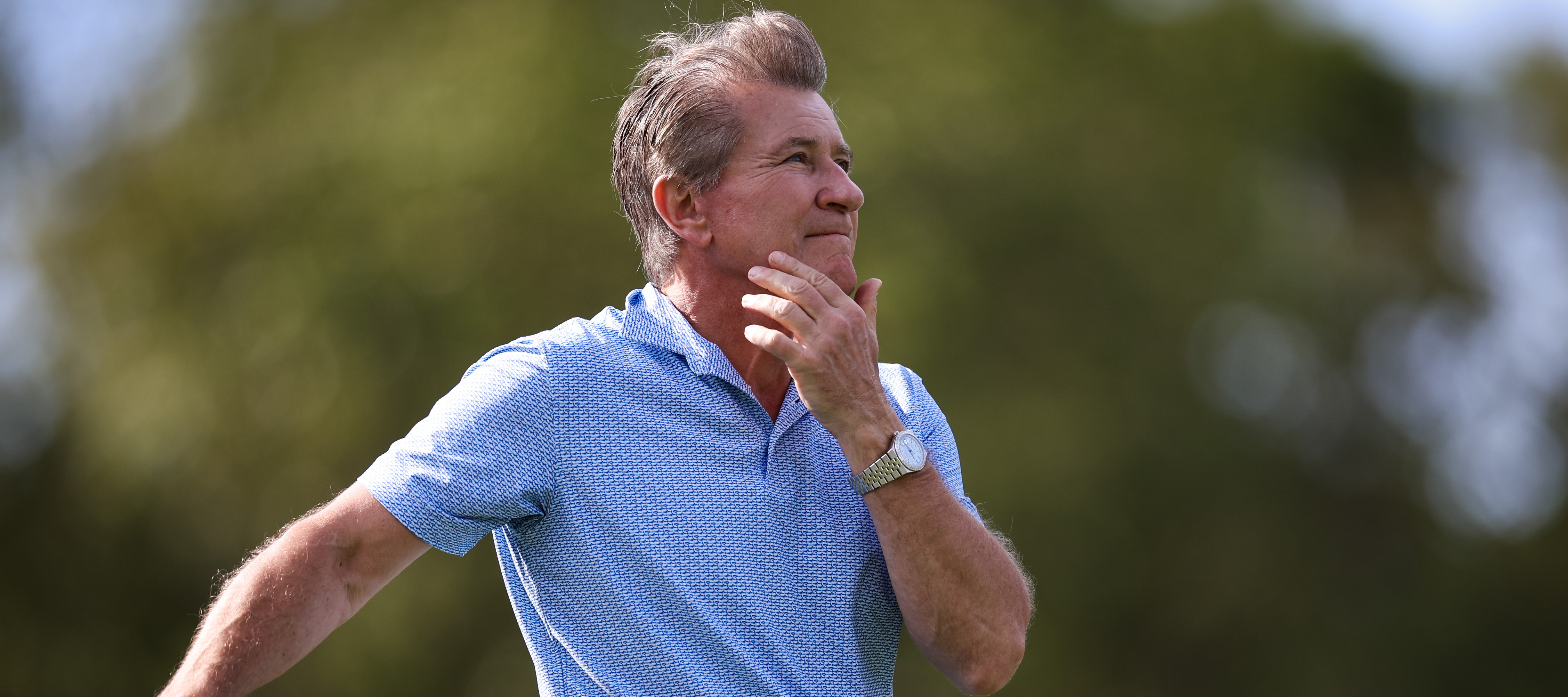A side profile shot of Robert Herjavec on a golf course, leaning on a club and holding his head in his hand.