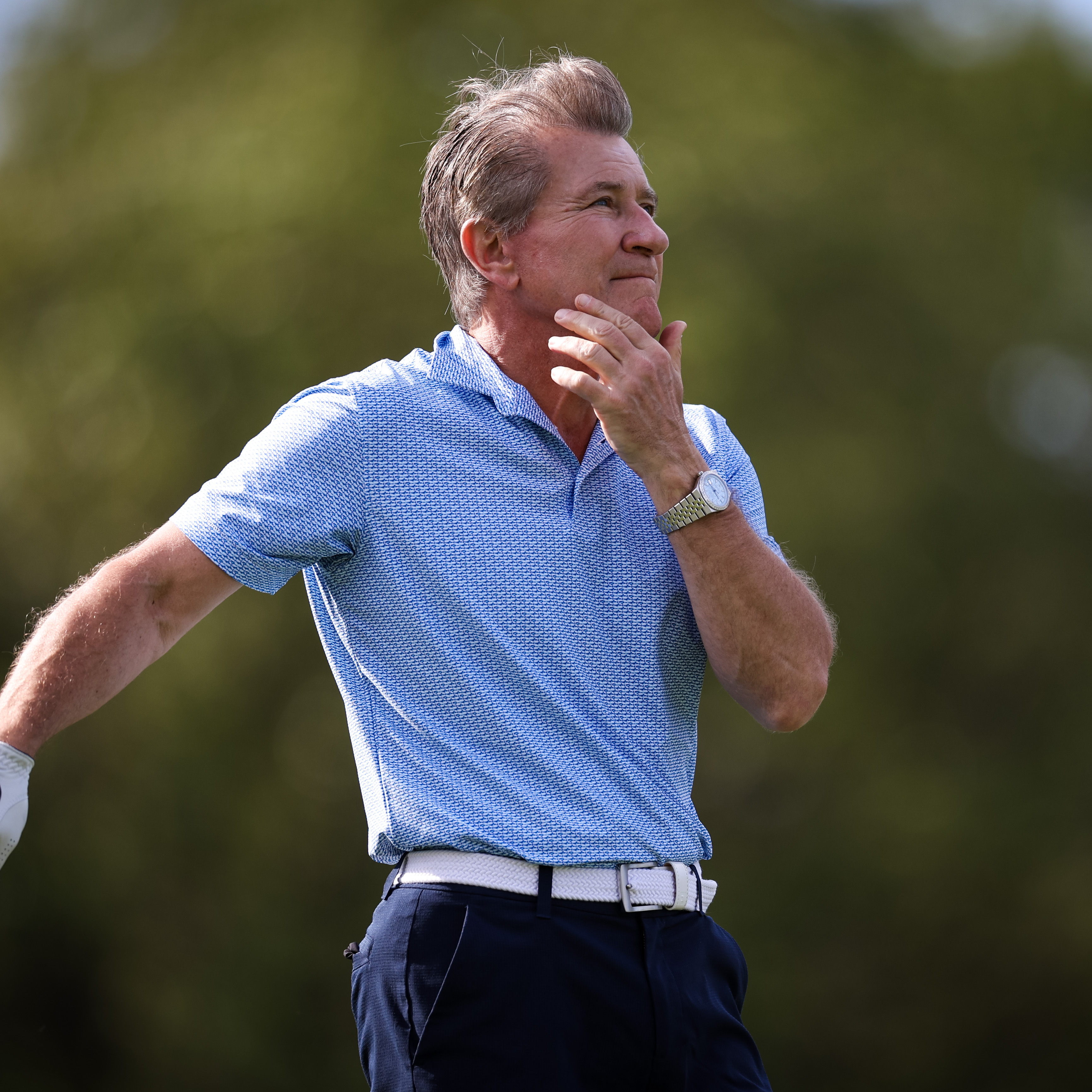 A side profile shot of Robert Herjavec on a golf course, leaning on a club and holding his head in his hand.
