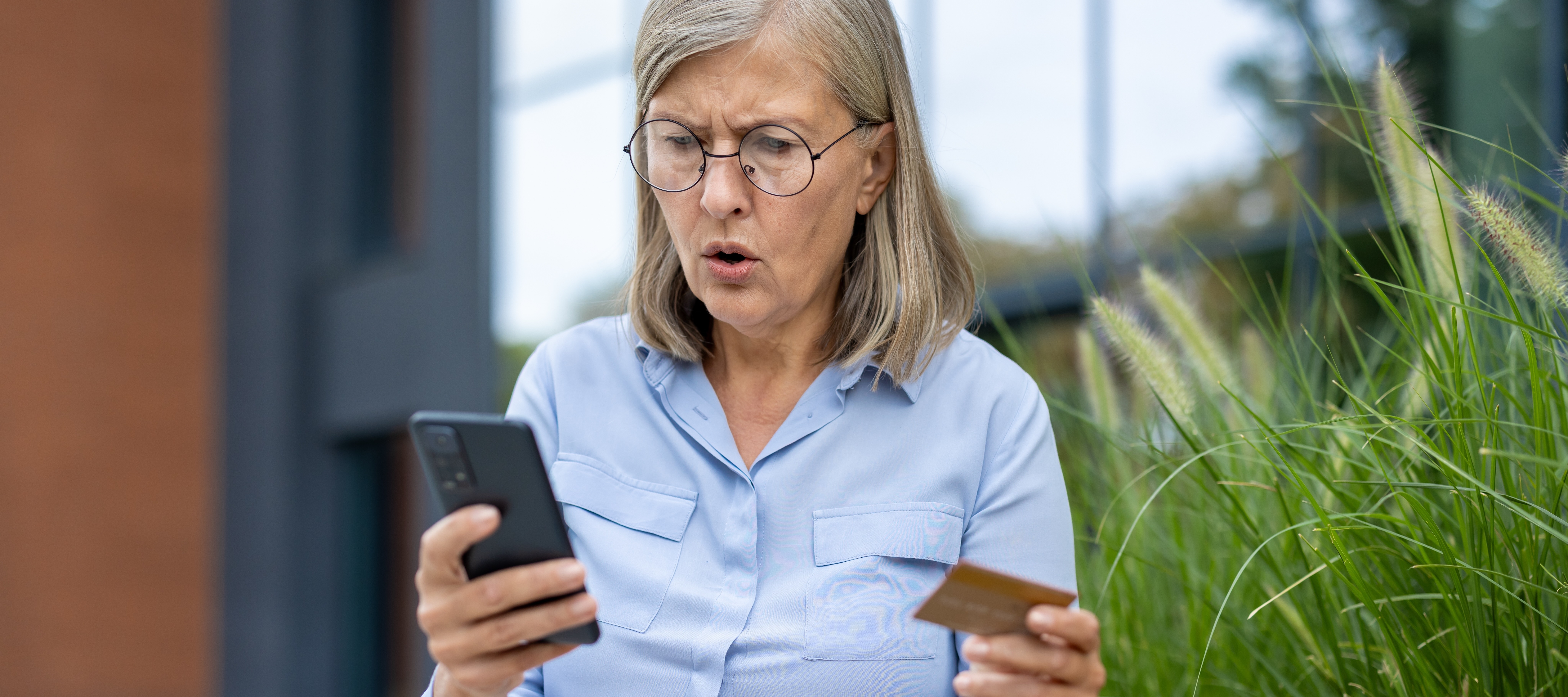 An older woman looks shocked while holding a credit card and smartphone outdoors, likely experiencing a problem.
