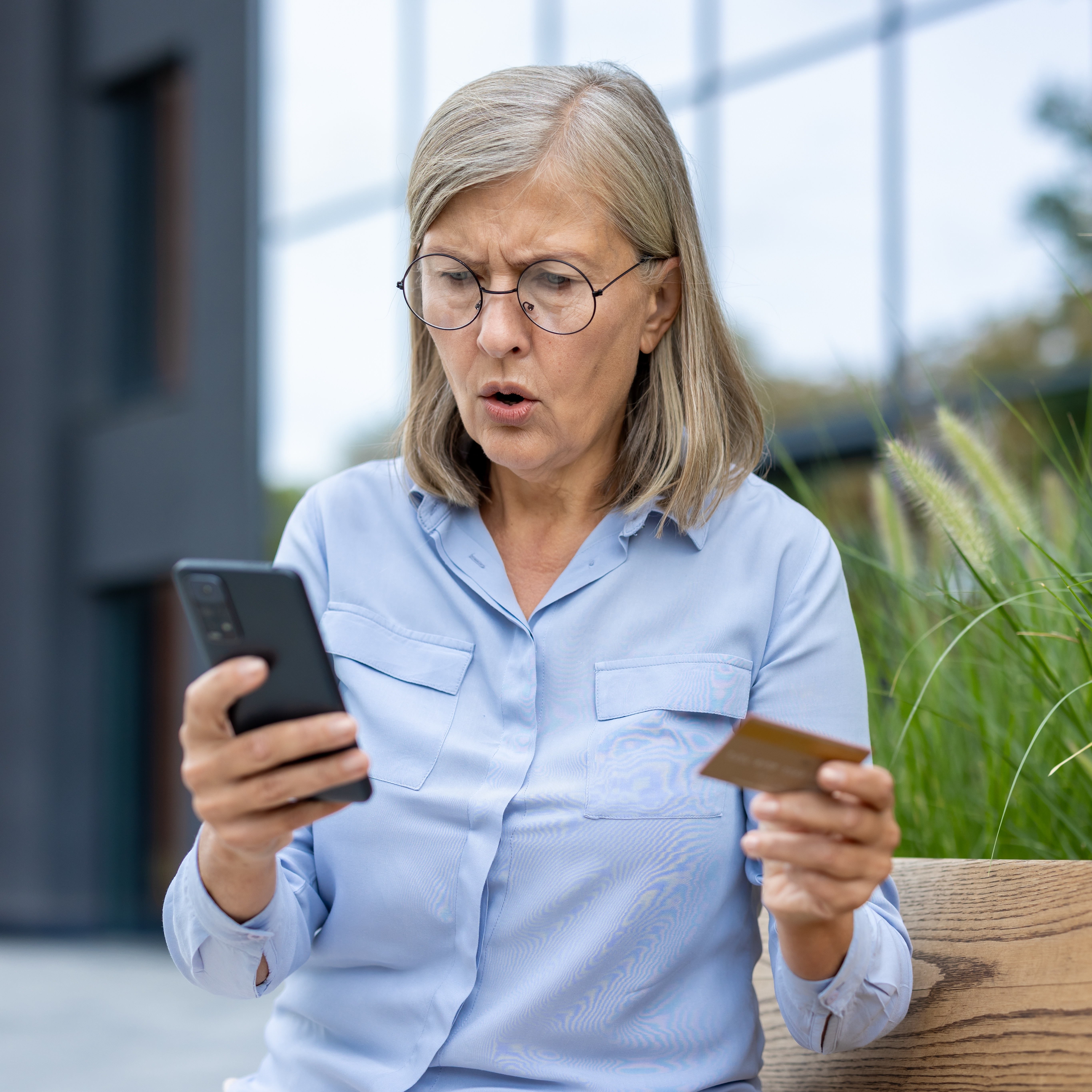 An older woman looks shocked while holding a credit card and smartphone outdoors, likely experiencing a problem.