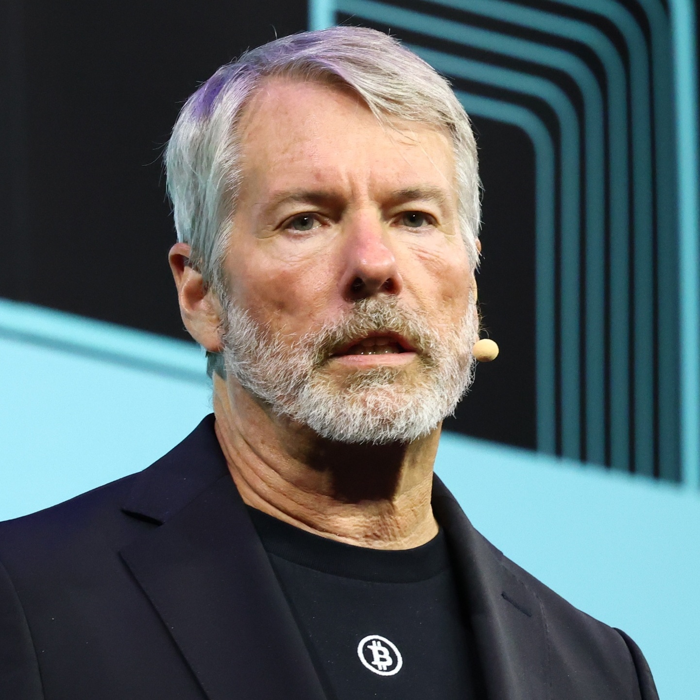 Michael J. Saylor speaks onstage at Consensus Miami 2026. A text bubble is overlayed, showing an X post from his account, saying "Never sell your Bitcoin."