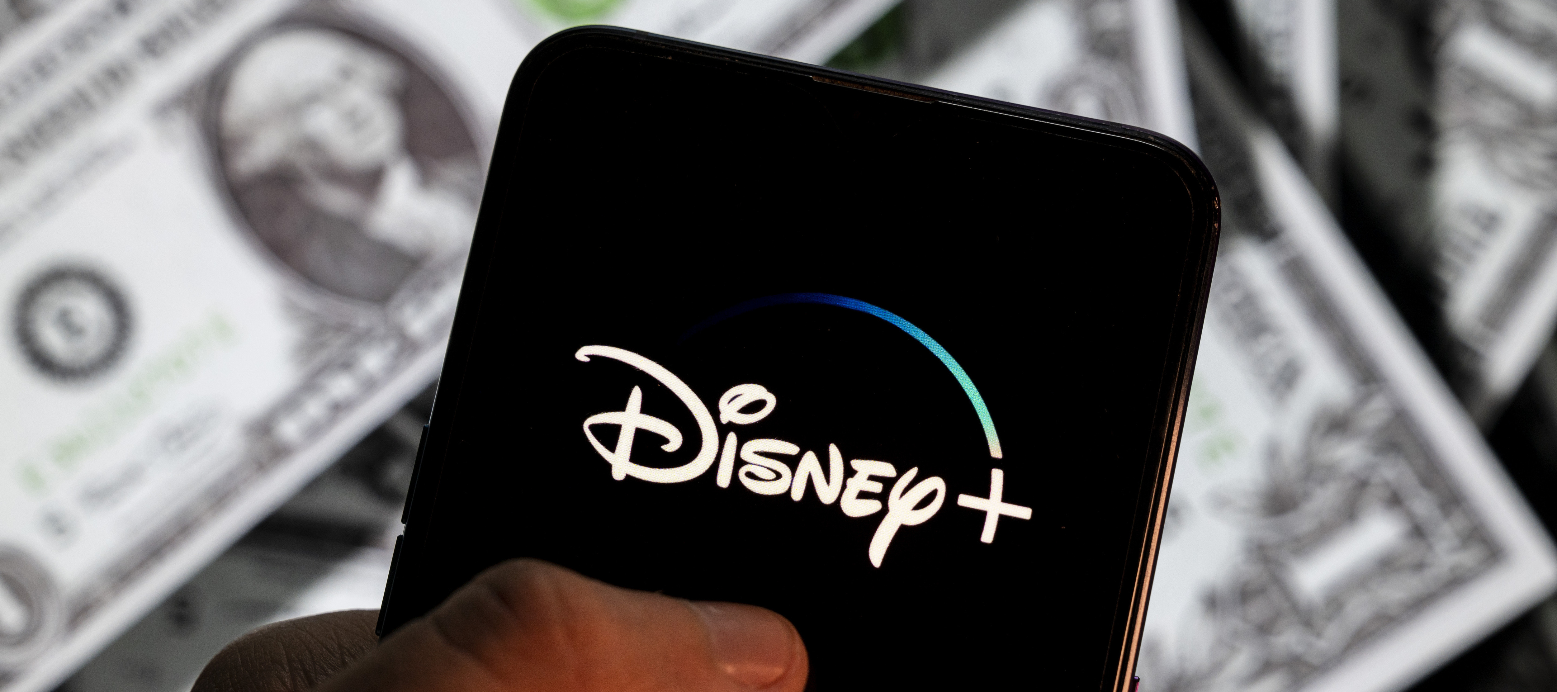 The Disney Plus logo is shown on a smartphone that is held over a pile of money.