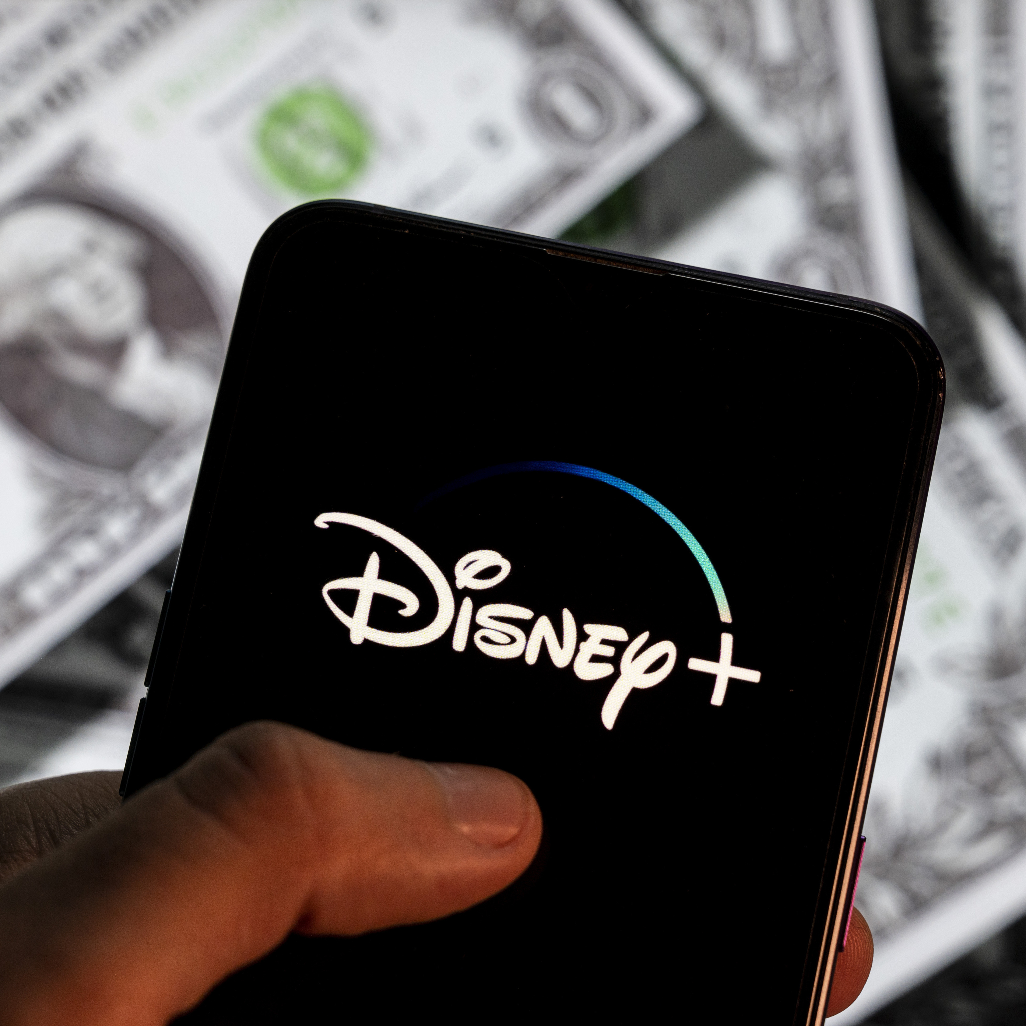The Disney Plus logo is shown on a smartphone that is held over a pile of money.