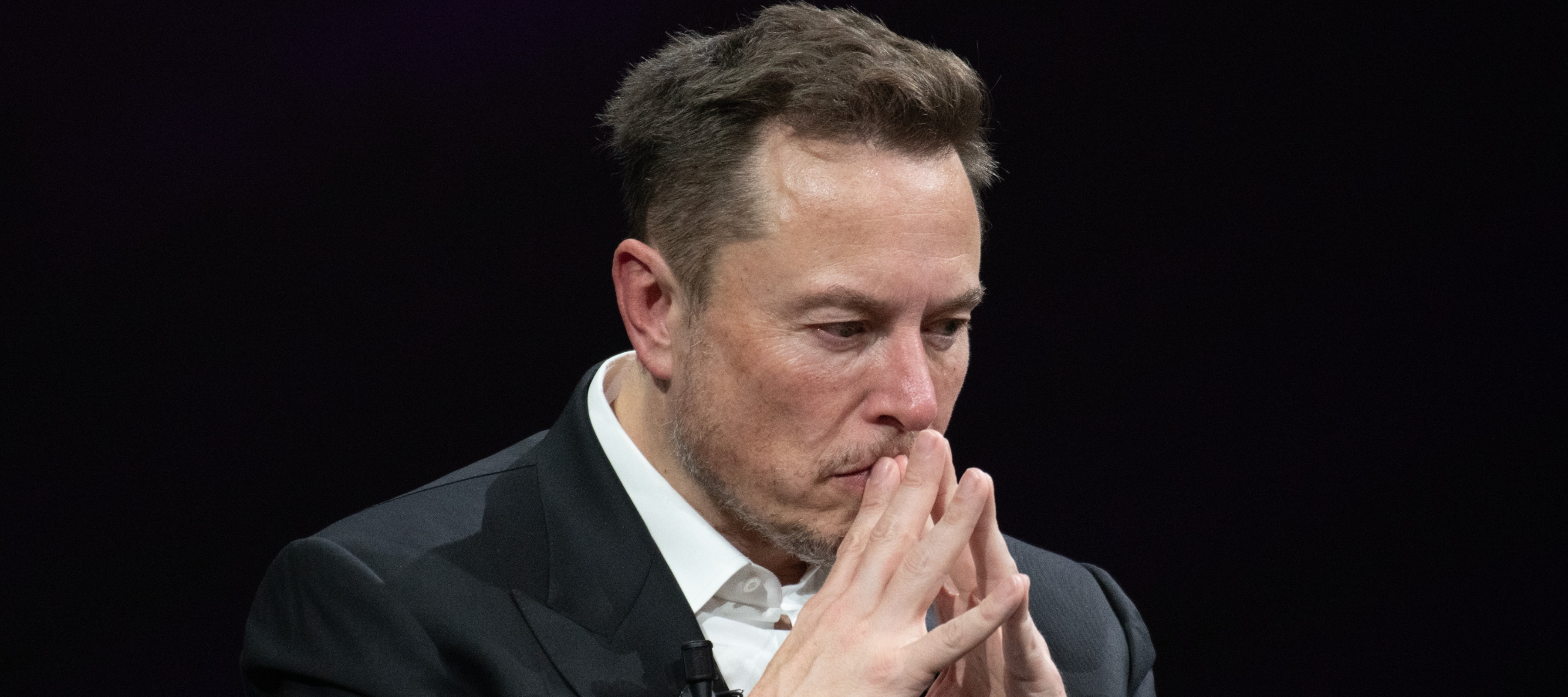 Elon Musk looking concerned with his hands folded near his face