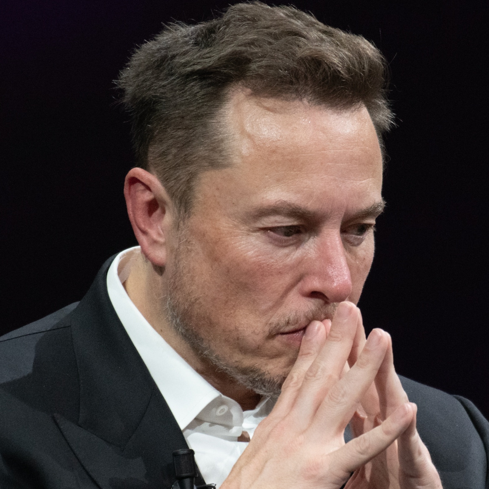 Elon Musk looking concerned with his hands folded near his face