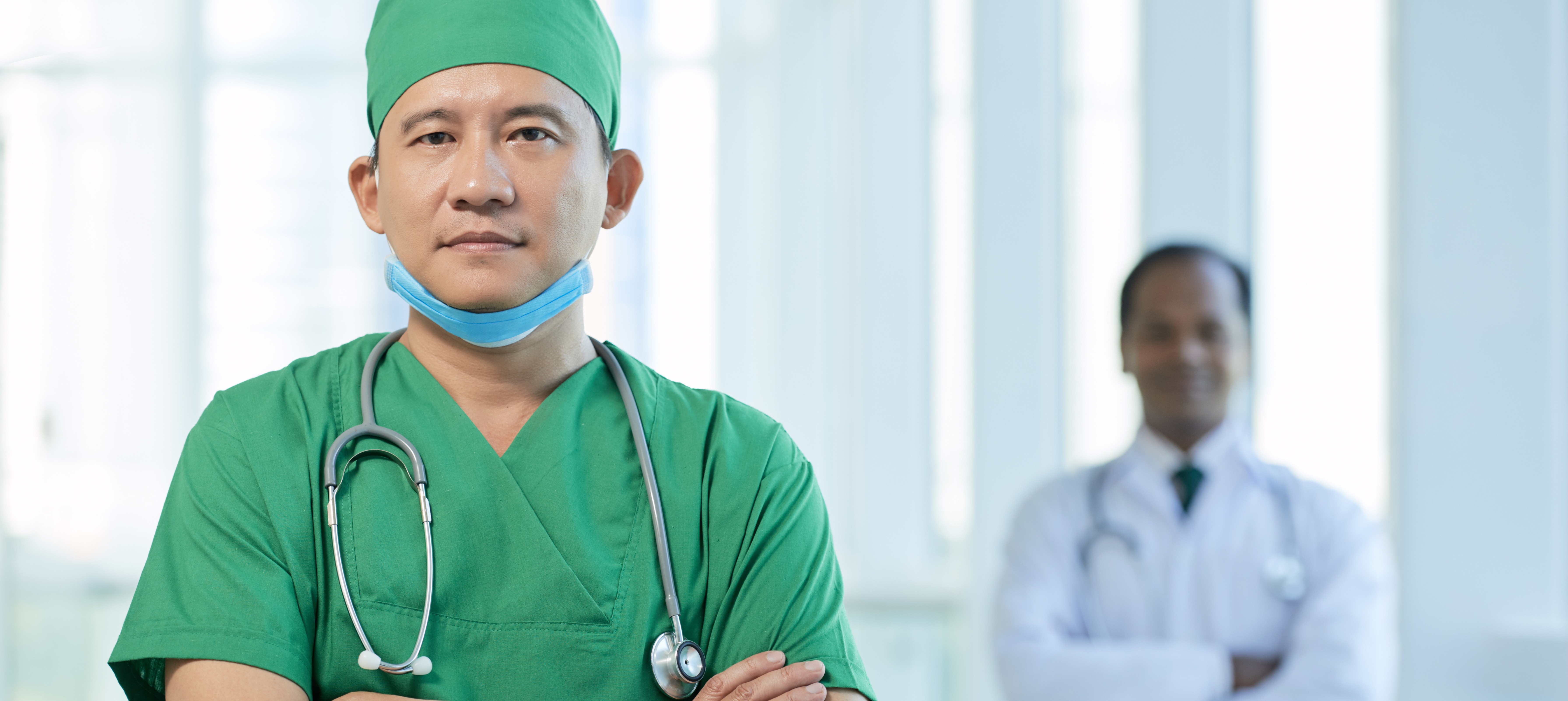 A man wearing green scrubs and a matching surgical cap stands in a hospital hallway. Behind him, out of focus, a person in a white coat stands with their arms crossed as well.