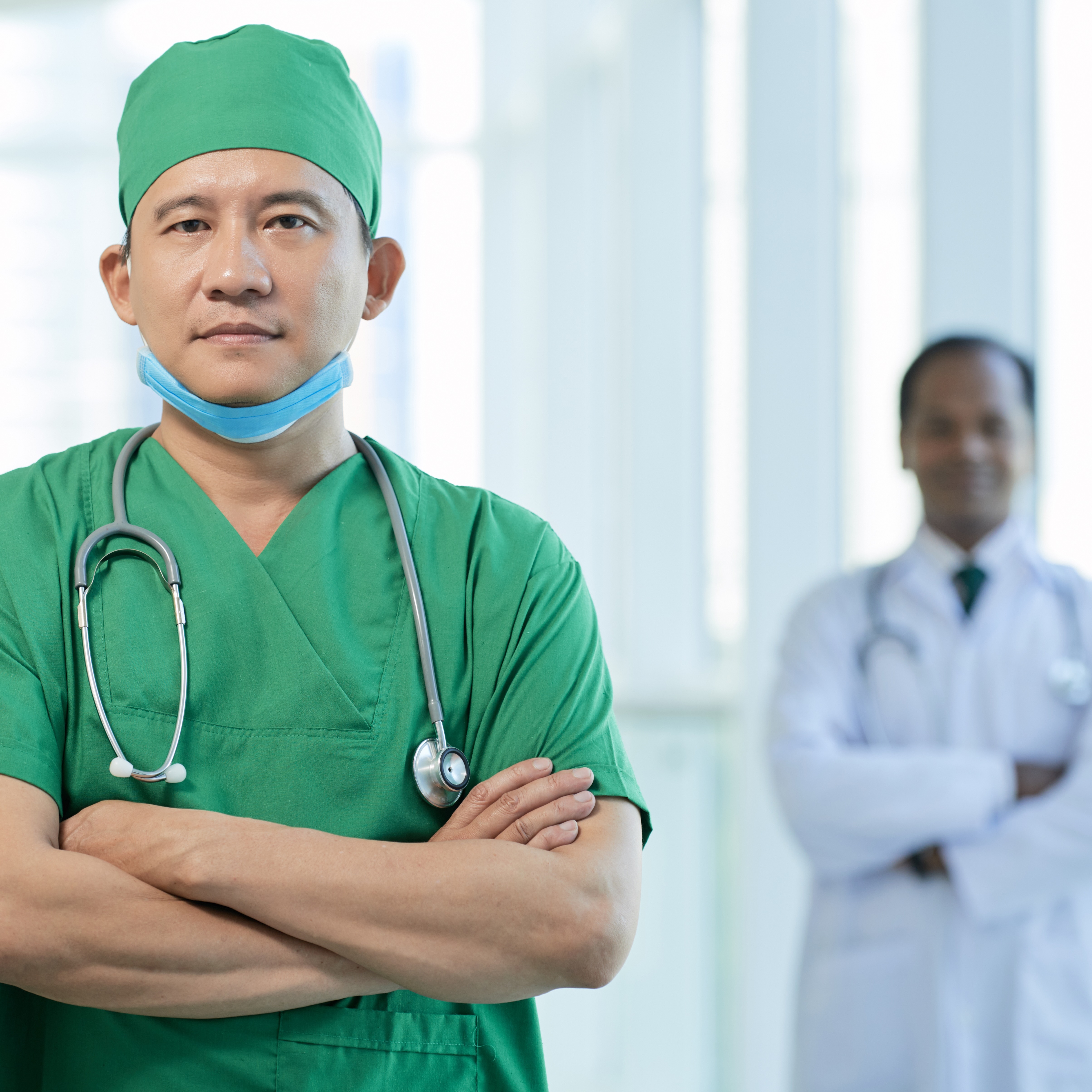 A man wearing green scrubs and a matching surgical cap stands in a hospital hallway. Behind him, out of focus, a person in a white coat stands with their arms crossed as well.