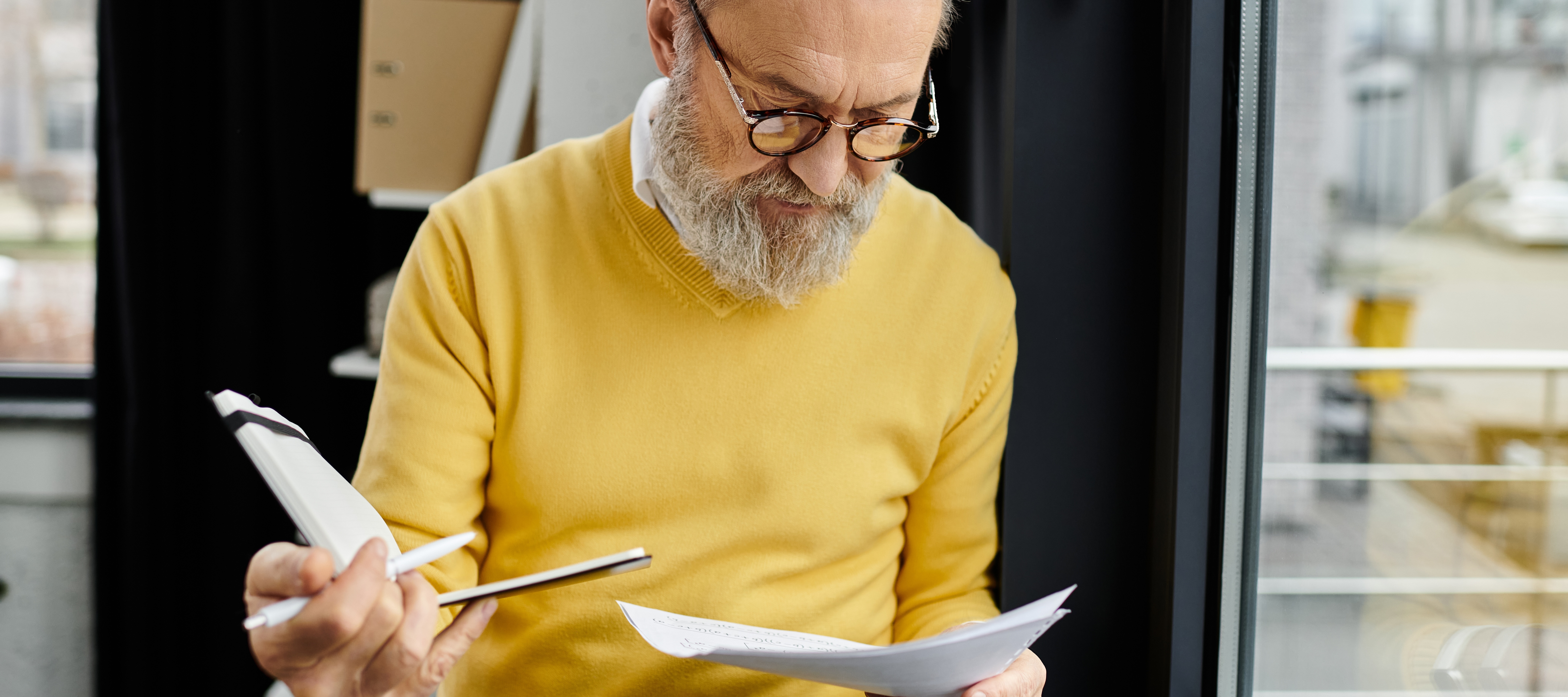 A man with grey hair wearing a yellow sweater stands in front of a window, looking down at papers he is holding in his hands.