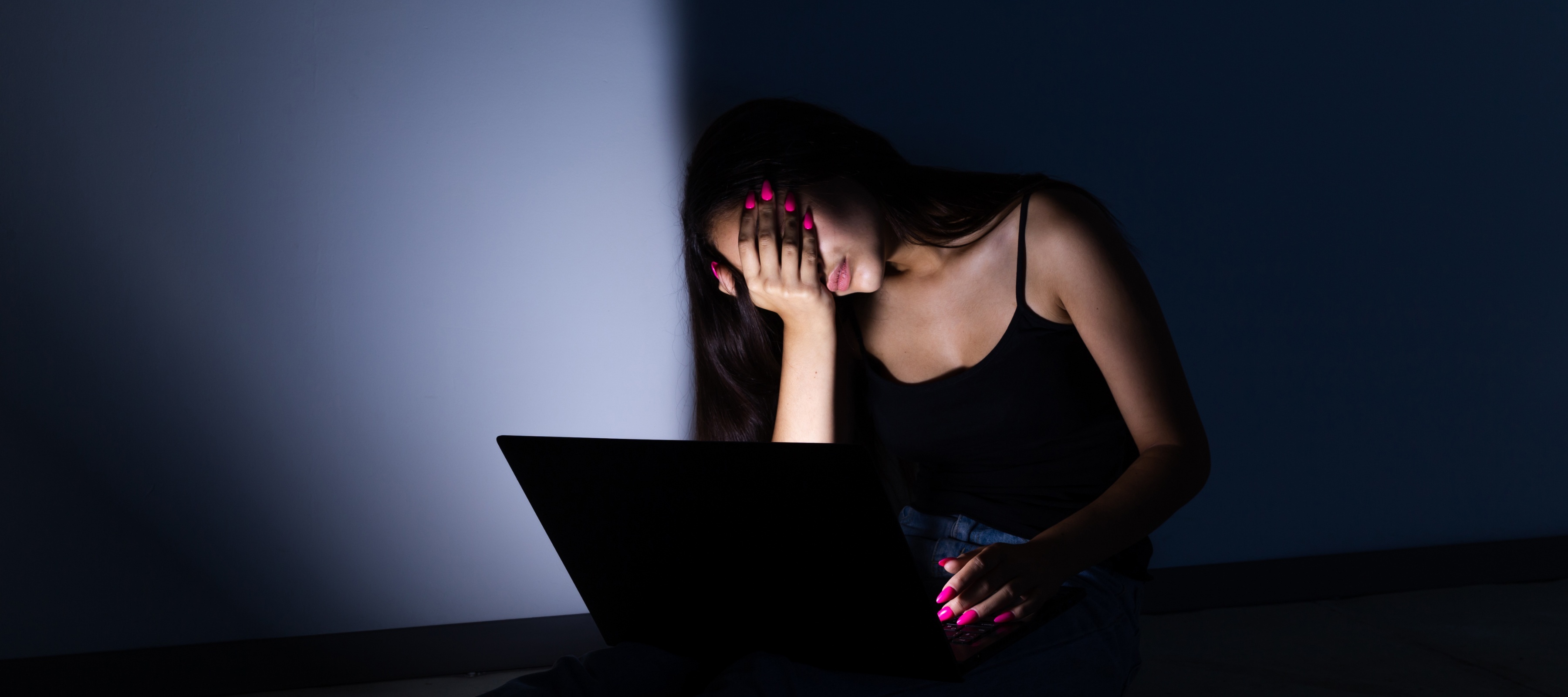 A young woman in front of a laptop, in the dark, with her hand covering her face
