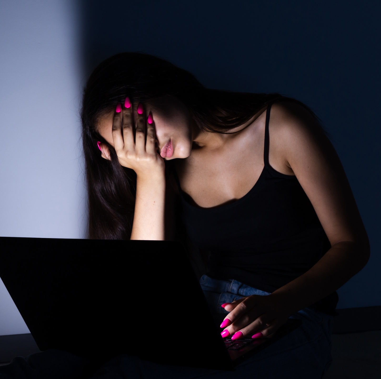 A young woman in front of a laptop, in the dark, with her hand covering her face