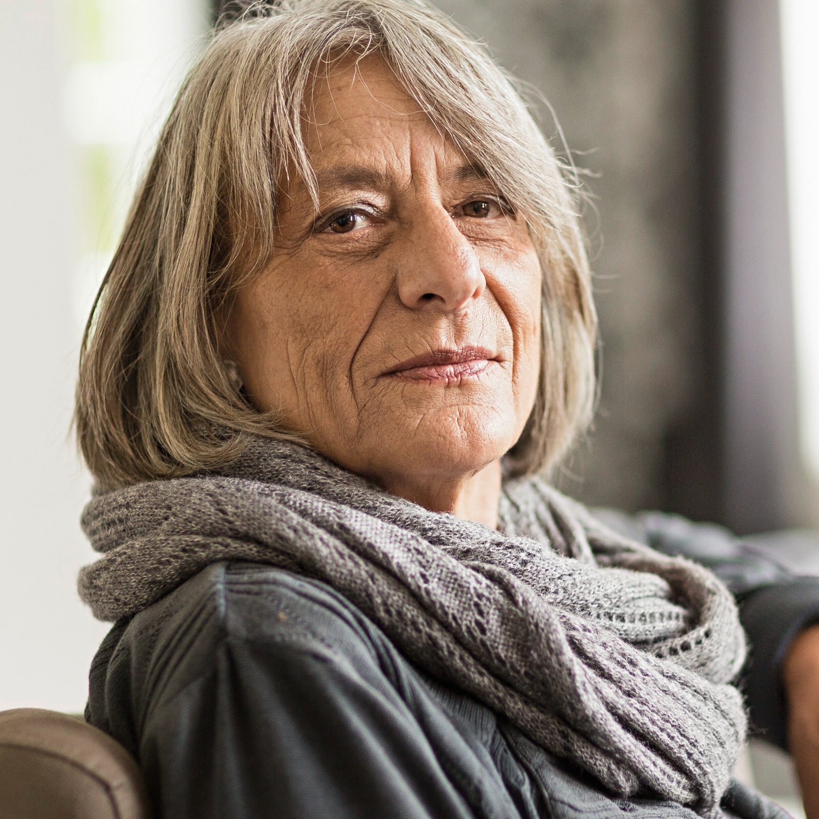 Older woman sitting in a chair, looking directly at the camera with a neutral, interested face.