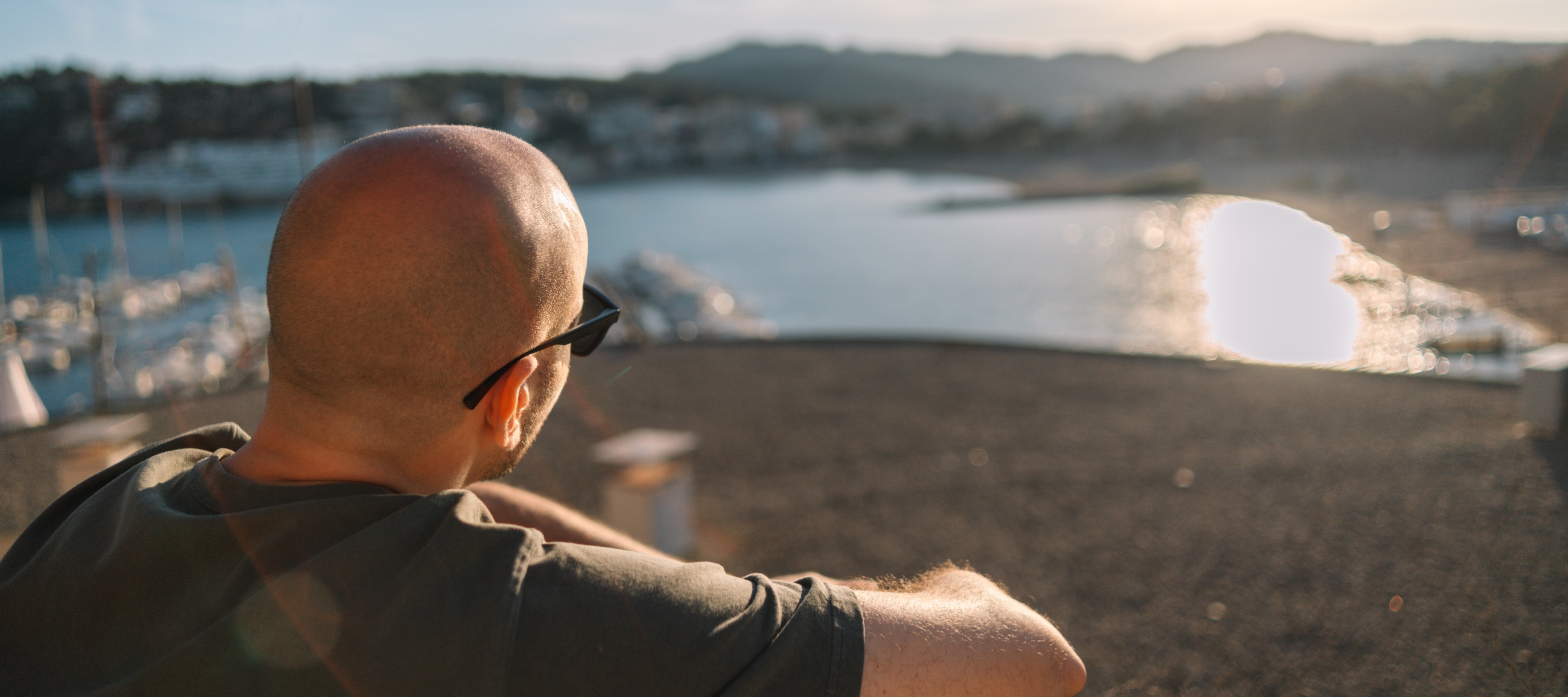 A bald man in glasses sitting at the beach overlooking a lake.